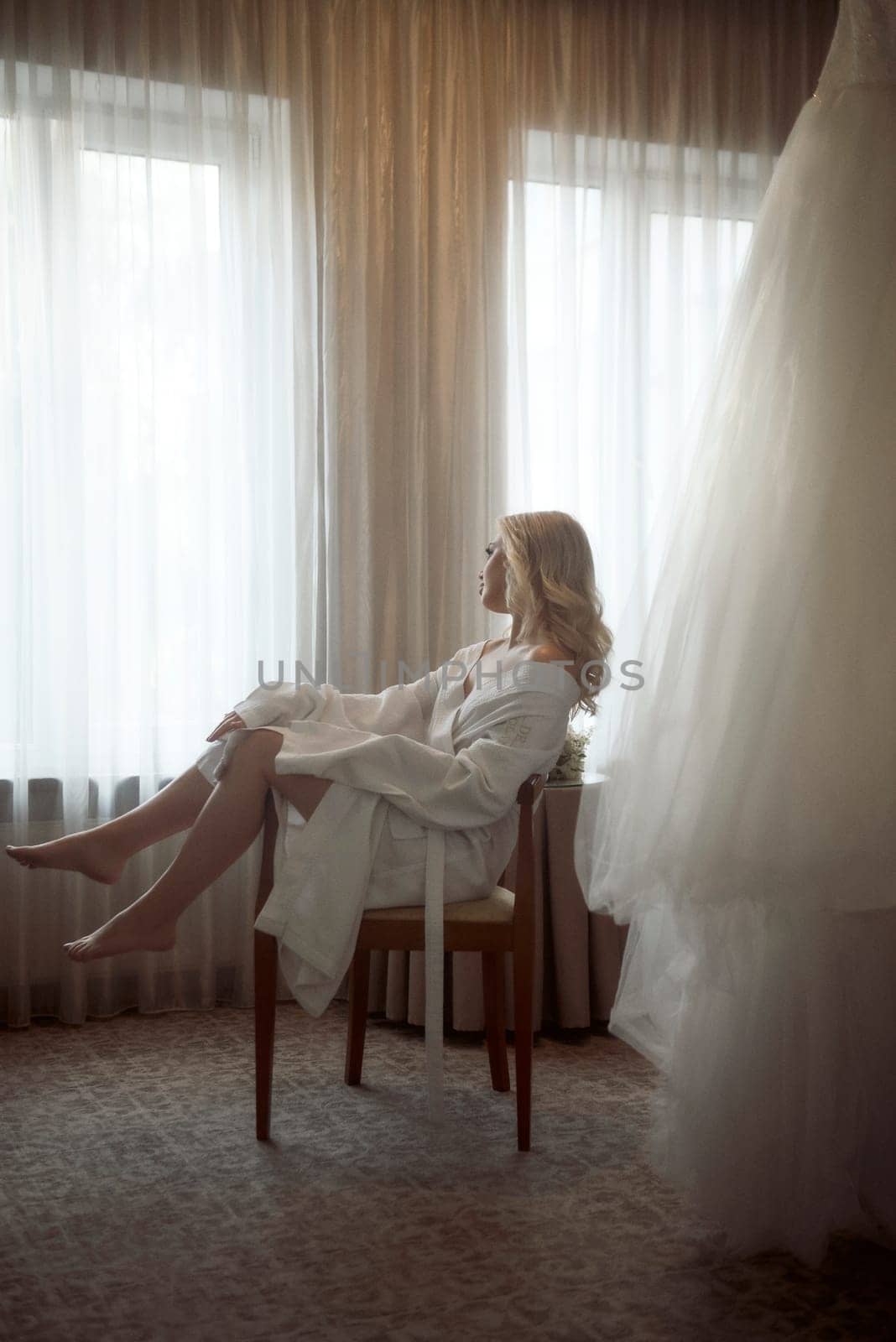 beautiful girl bride sits in chair in hotel on wedding morning. blonde sits and dreams about wedding, with hairdo in white robe, getting ready for wedding day, wedding dress nearby