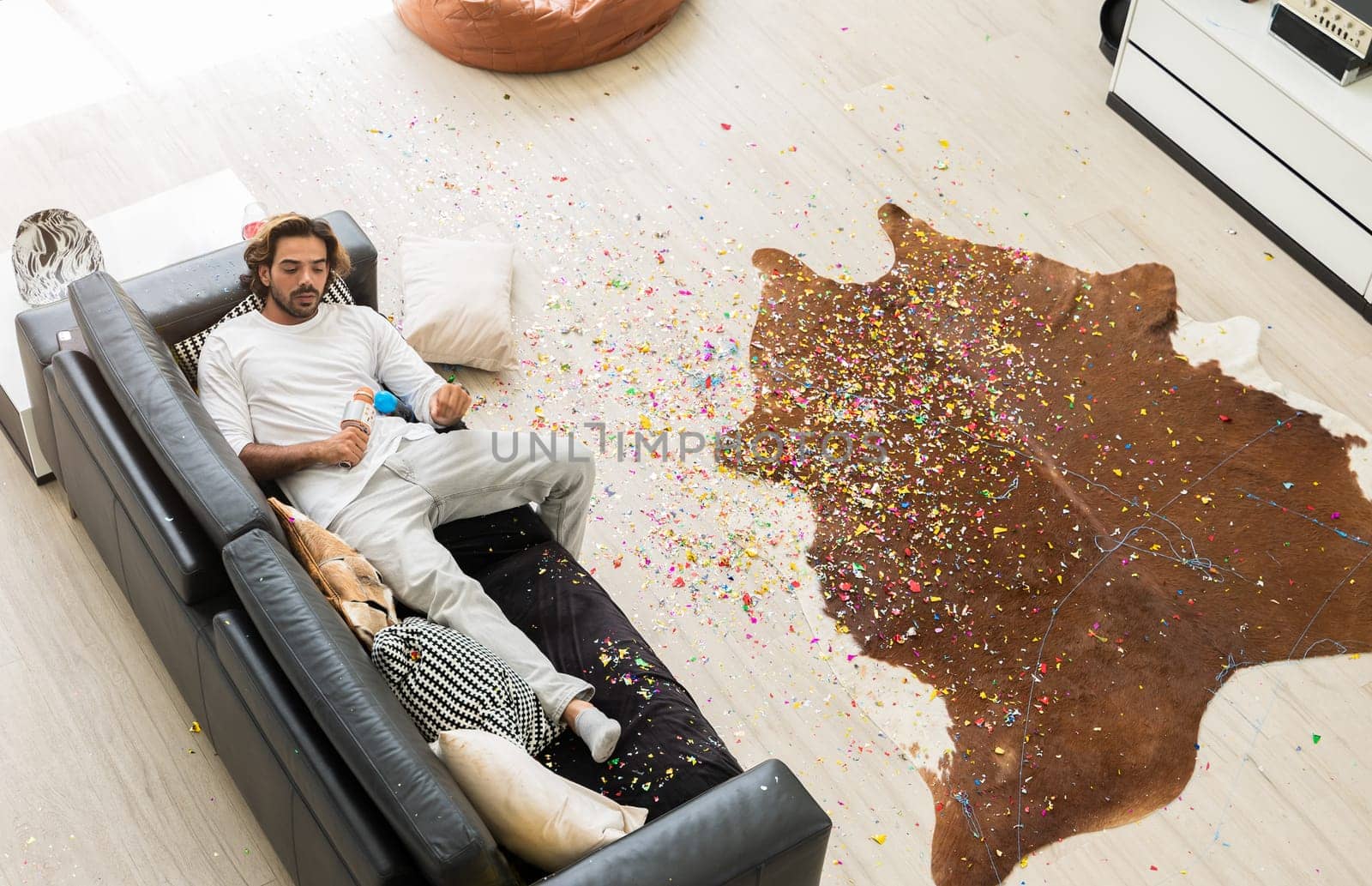 Man lying on black leather couch and holding microphone in disheveled room with colorful confetti on wooden floor after Christmas celebration party at home.