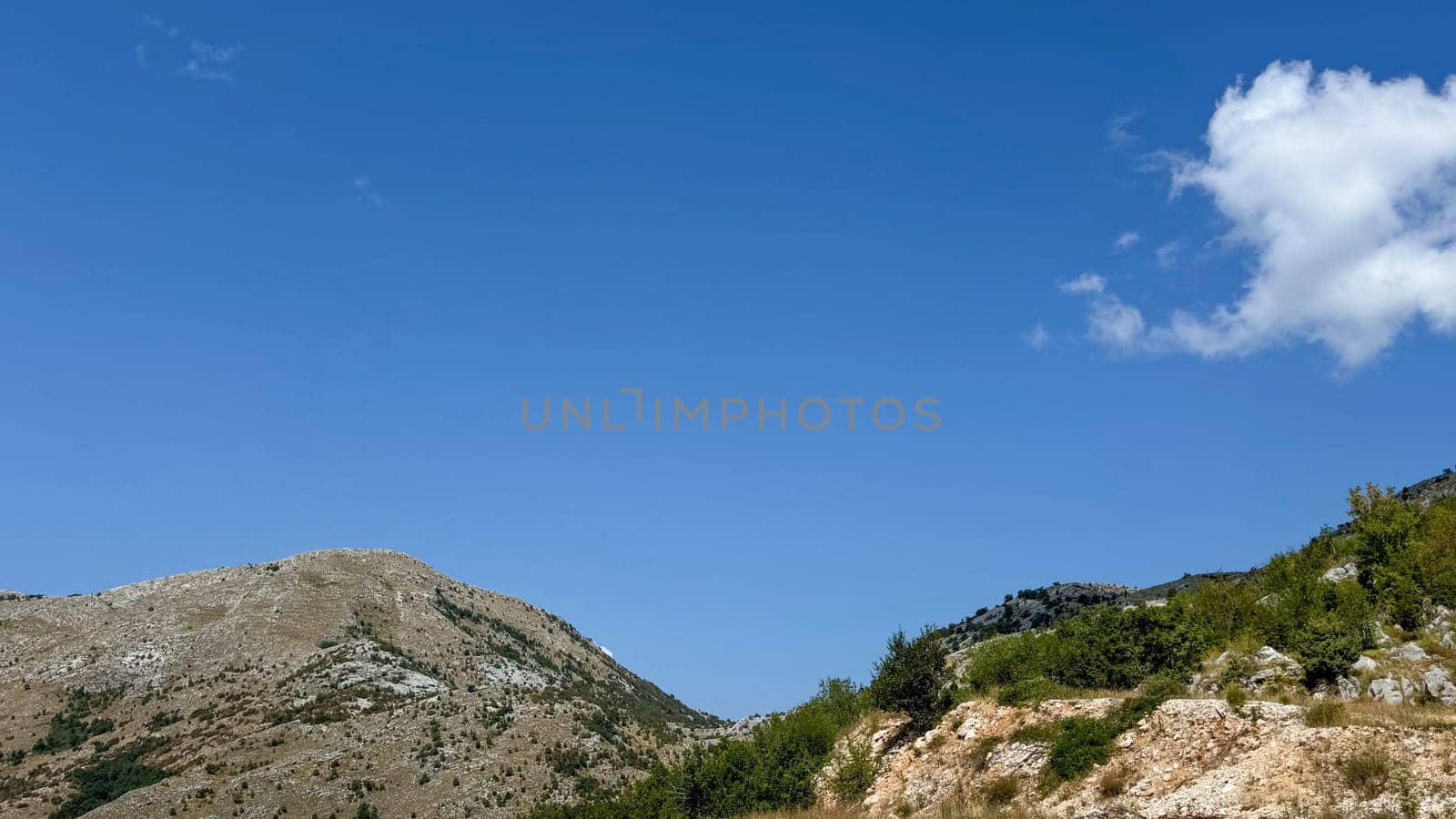 A minimalist landscape featuring a rocky hill with sparse vegetation under a bright, clear blue sky.