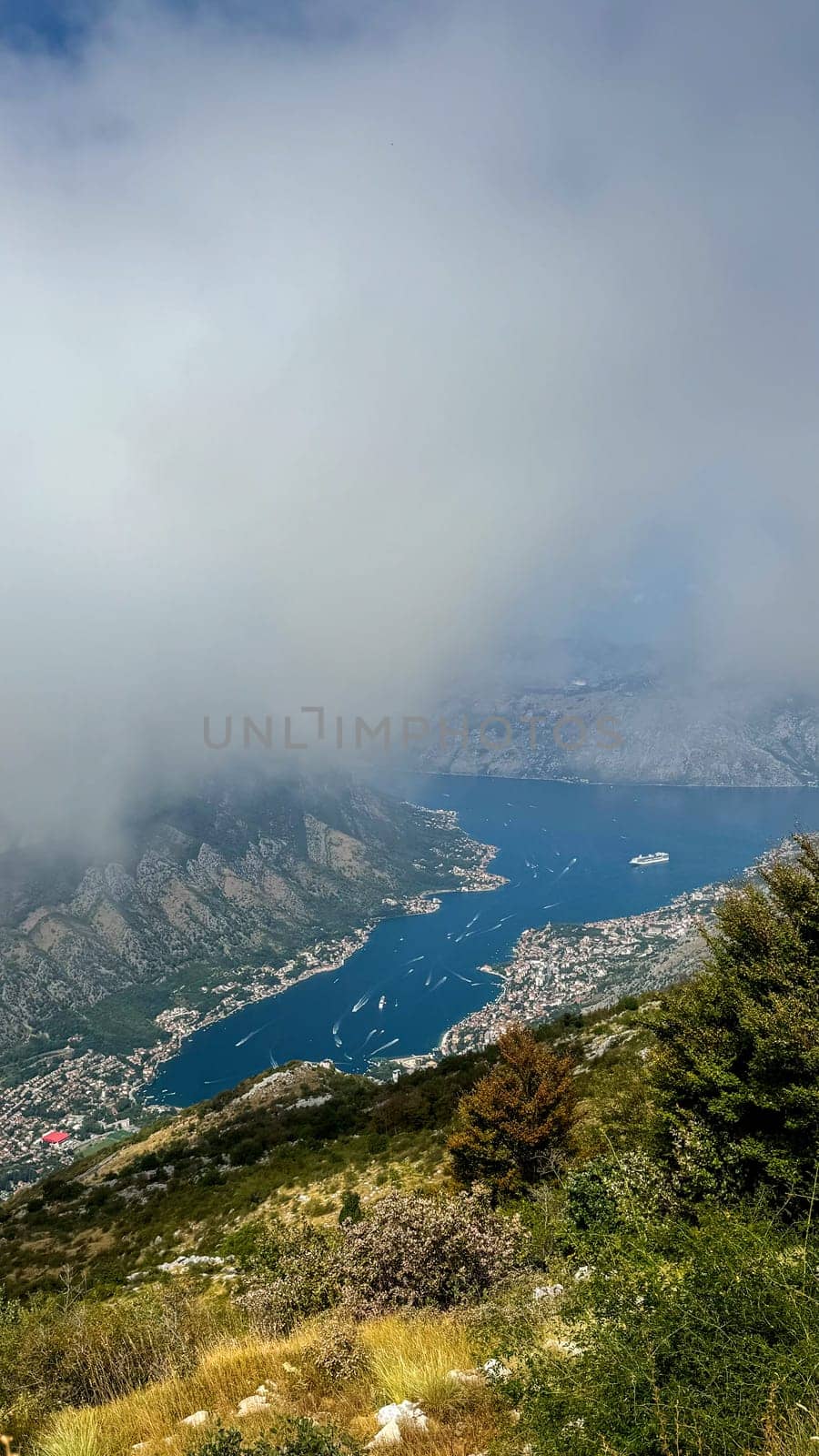 A misty aerial view of a coastal town nestled by a deep blue bay, partially obscured by low-hanging clouds.