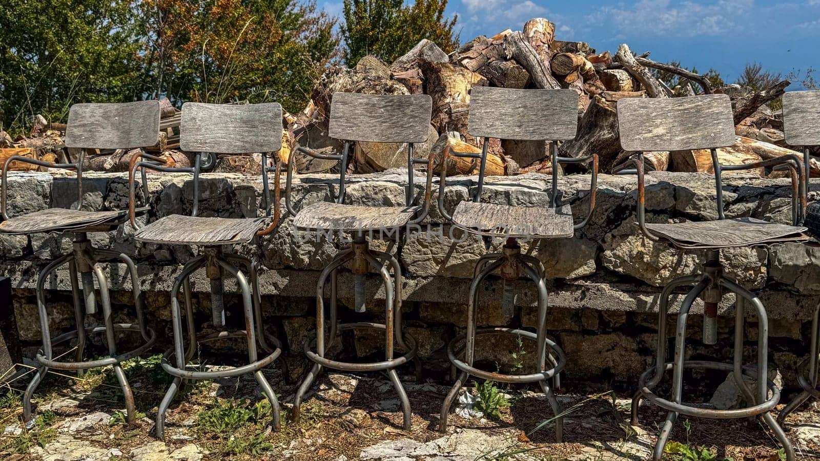 A close-up of six wooden bar stools with rusty metal frames, lined up against a stone wall, against a background of chopped wood and green trees under a clear sky.decay and outdoor decor themes.