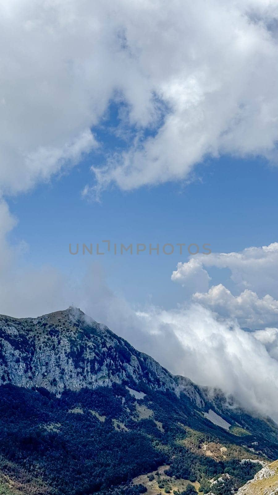 A dramatic scene of a mountain peak covered in dense greenery, partially hidden by swirling clouds under a bright blue sky.