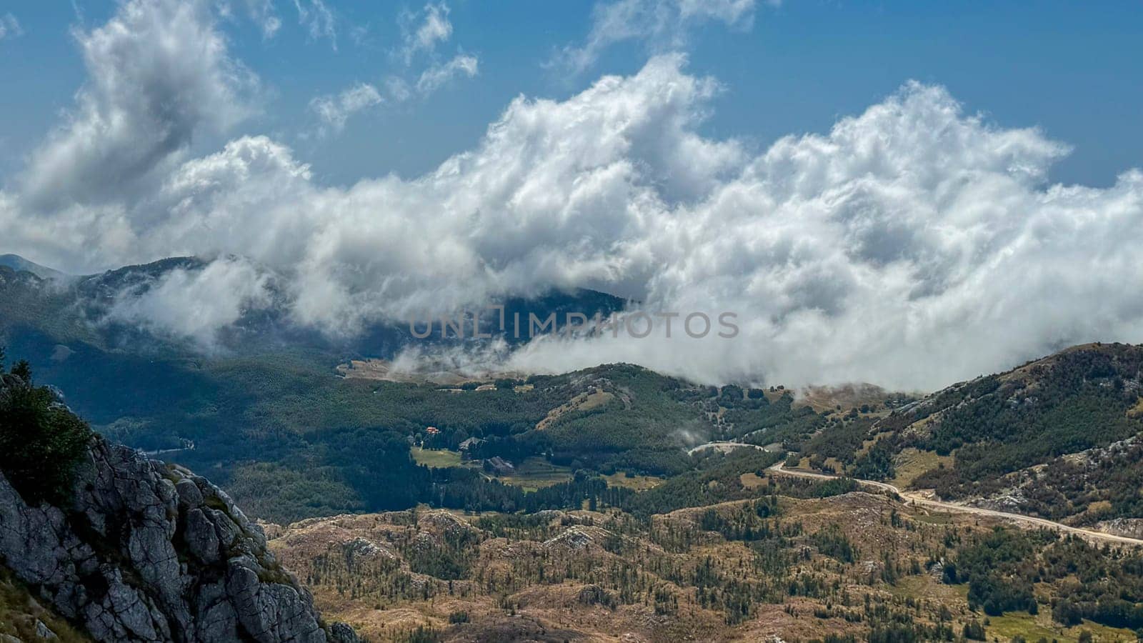 Scenic scene of low clouds rolling over green valleys and hills, with rugged mountains in the background. The landscape is bathed in sunlight under a clear sky.