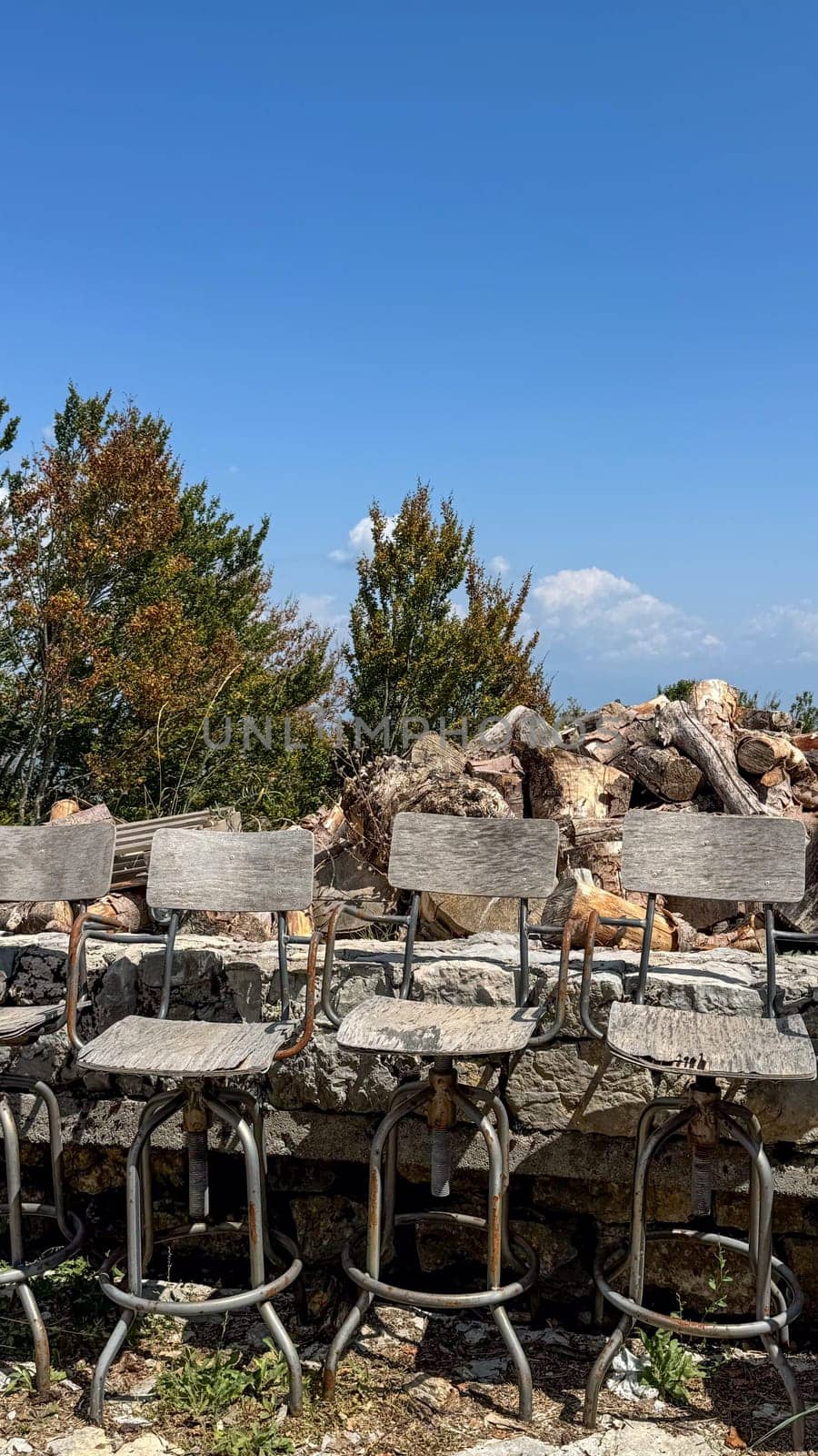 Old weathered wooden high bar stools with rusty metal frames against a background of green trees, piles of firewood and clear blue sky.