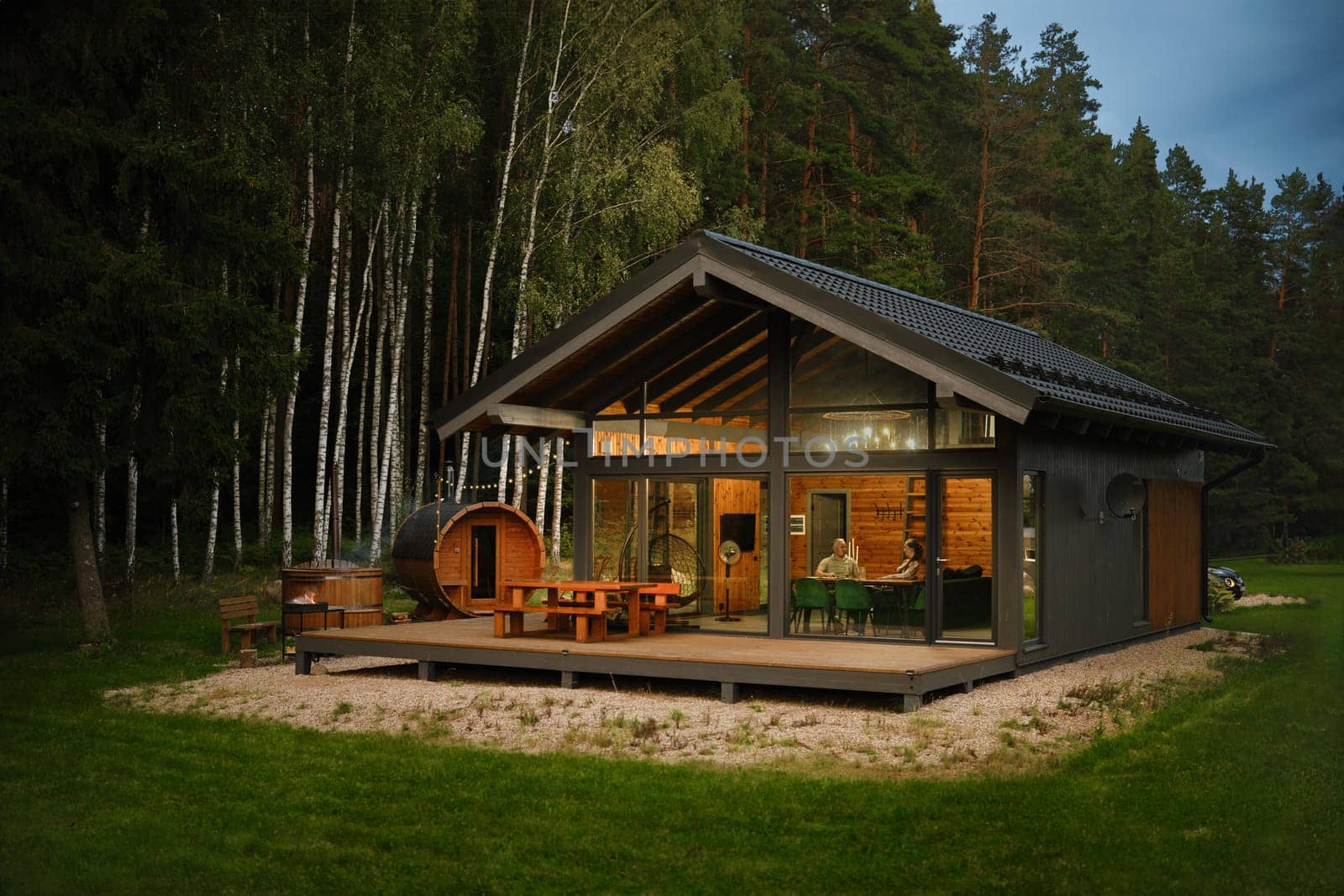 A modern cabin with a large glass windows and wooden deck with patio furniture at a forest clearing and a barrel-shaped wooden sauna