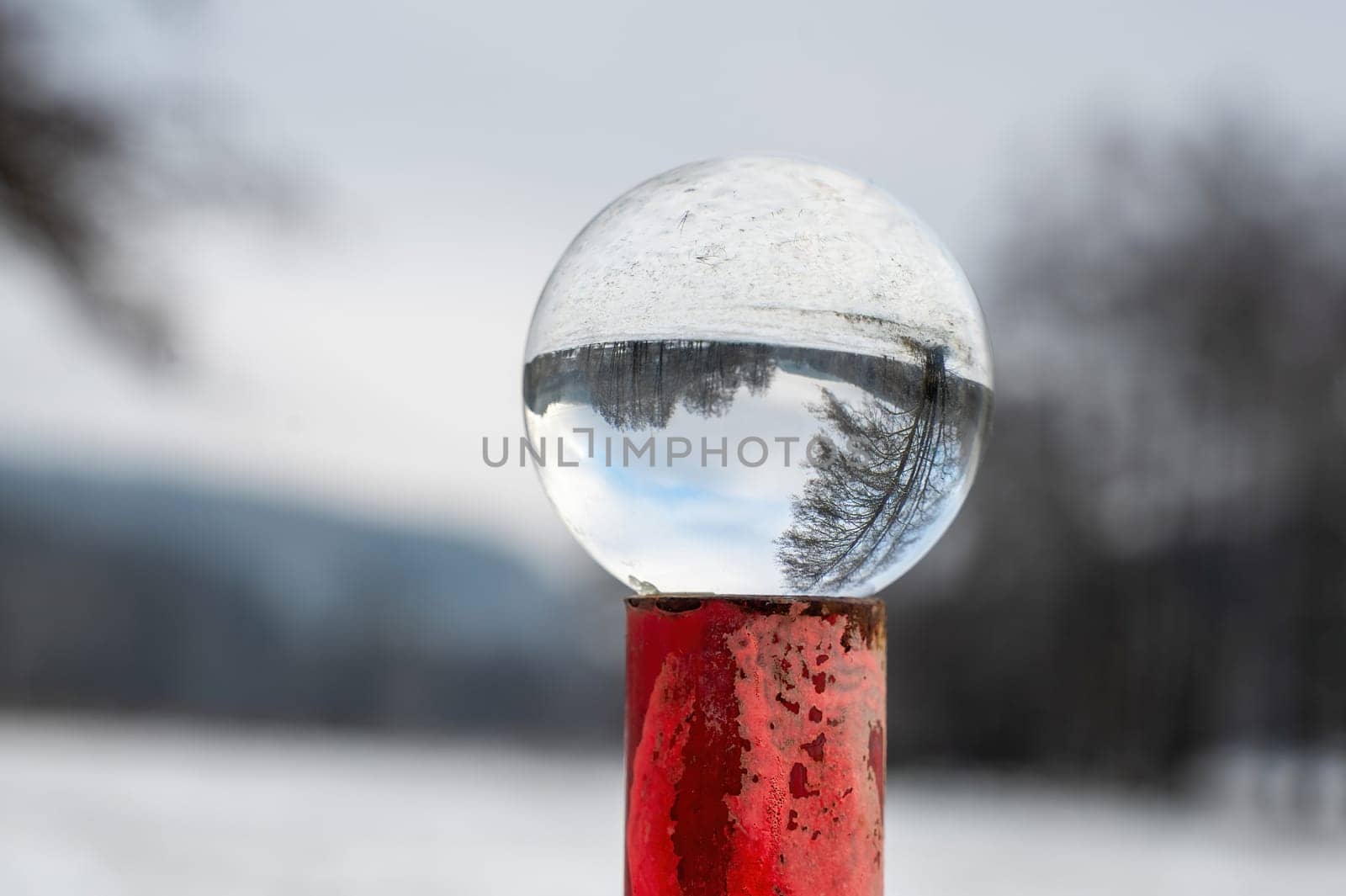 Glass ball lying on a rusty red metal post on a cold winter day in nature, with reflections of a snowy landscape in the glass