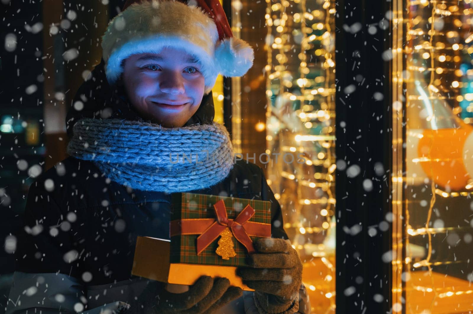 Boy in Santas hat with gifts box near illuminated shop window. Xmas presents holidays, or shopping on New Year or Christmas concept