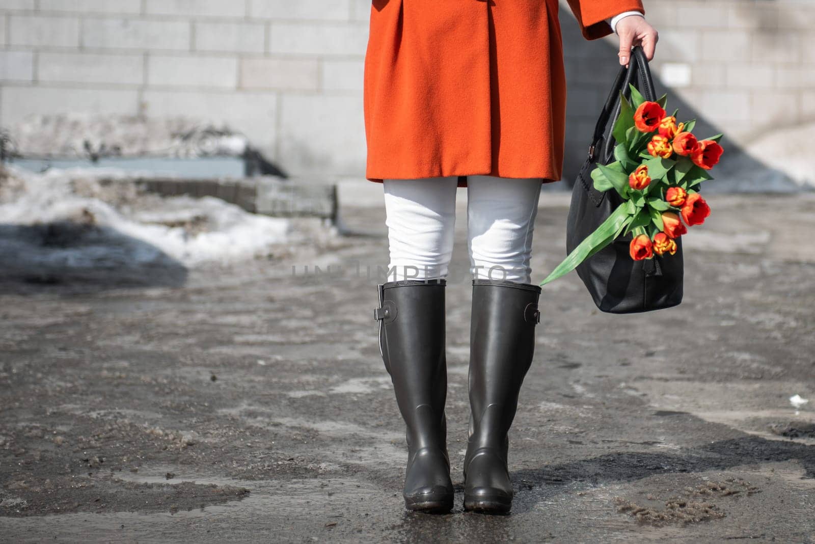Closeup of woman in a orange coat and rubber boots walking down the street with a bouquet of tulips in spring city
