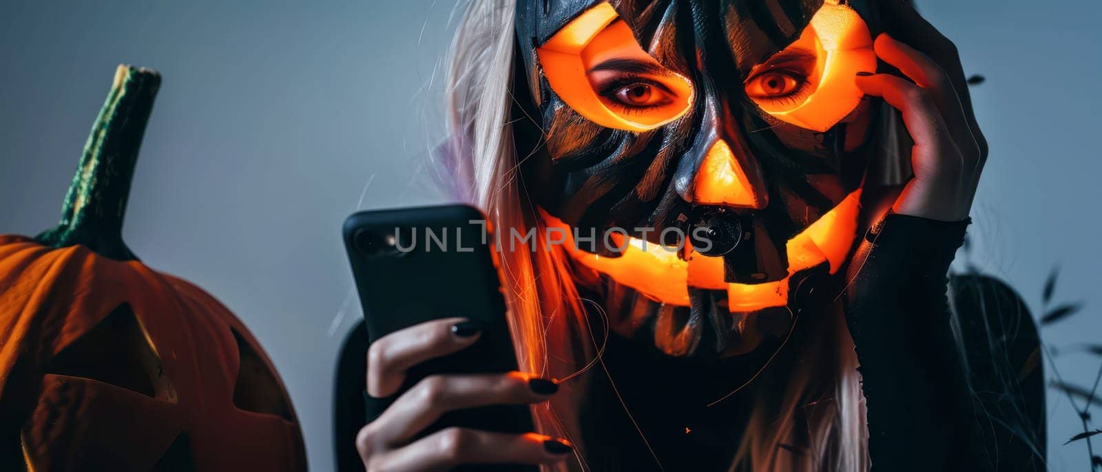 A person wears a creatively carved pumpkin mask while holding a smartphone. The atmosphere is festive, featuring pumpkins and dim lighting, enhancing the Halloween spirit.