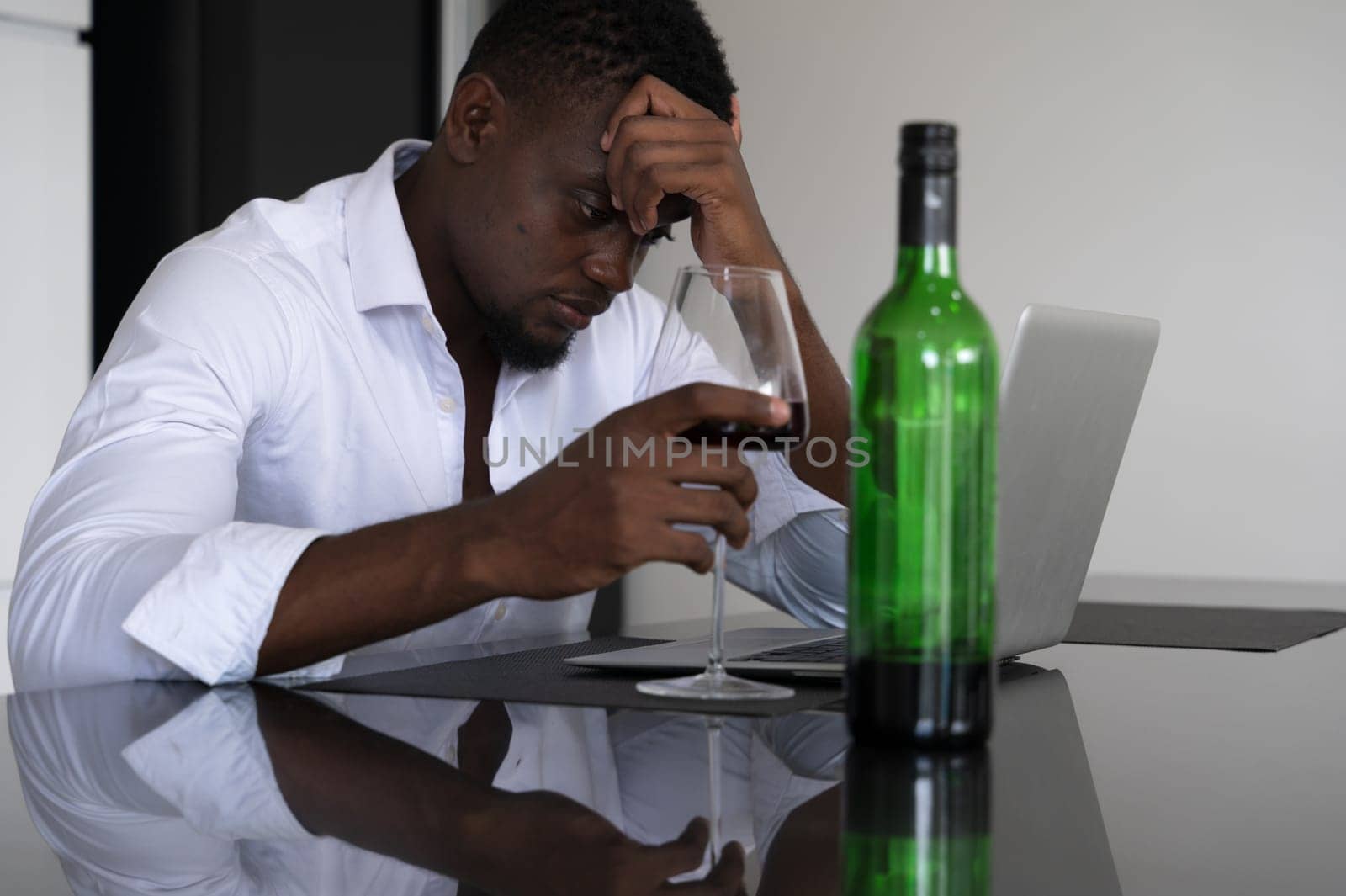 Businessman in white shirt sitting at kitchen tabletop holding head in one hand and glass of red wine in other. Black male appearing stressed or contemplative while working on laptop late night at home.