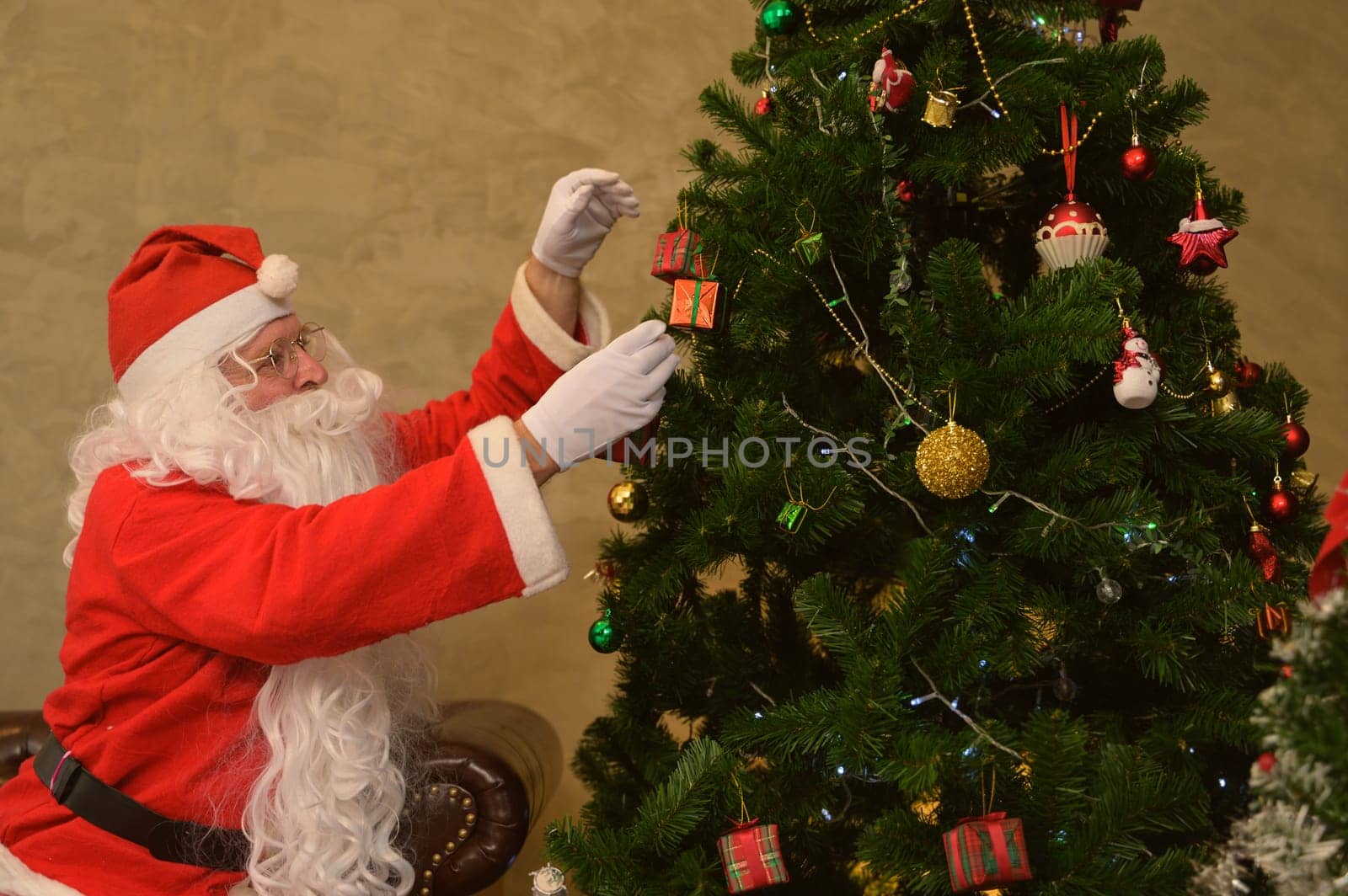 Happy Santa Claus in red suit decorating Christmas tree with balls, gift boxes and festive figurines in living room at home. Christmas and New Year concept.