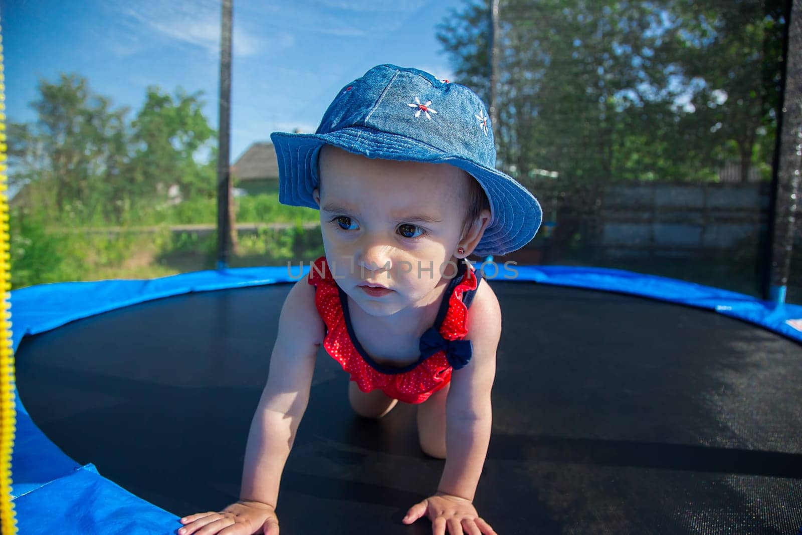 A small child in panama crawls on a large trampoline outdoors on a summer day