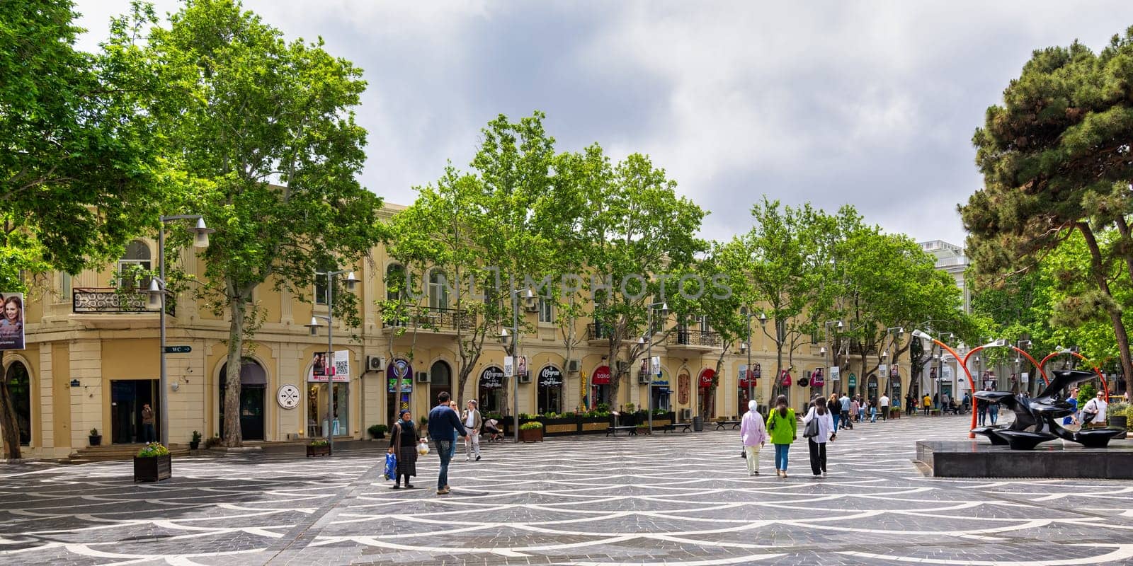 Baku, Azerbaijan - May 5, 2024: Visitors enjoy a leisurely walk down Nizami Street, surrounded by charming shops and lush trees under a cloudy sky, creating a lively urban atmosphere