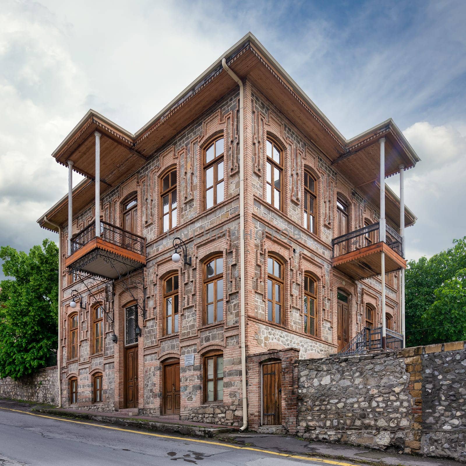 An elegant historic building stands in Shaki, Azerbaijan, featuring traditional architecture with detailed wooden balconies and large windows, against a backdrop of a cloudy sky.