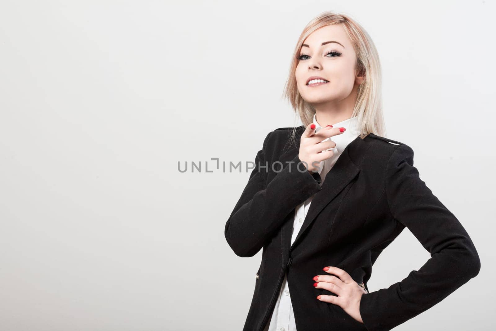 Young businesswoman is posing with her hand on her hip and pointing with her index finger. She is wearing a black jacket and a white shirt