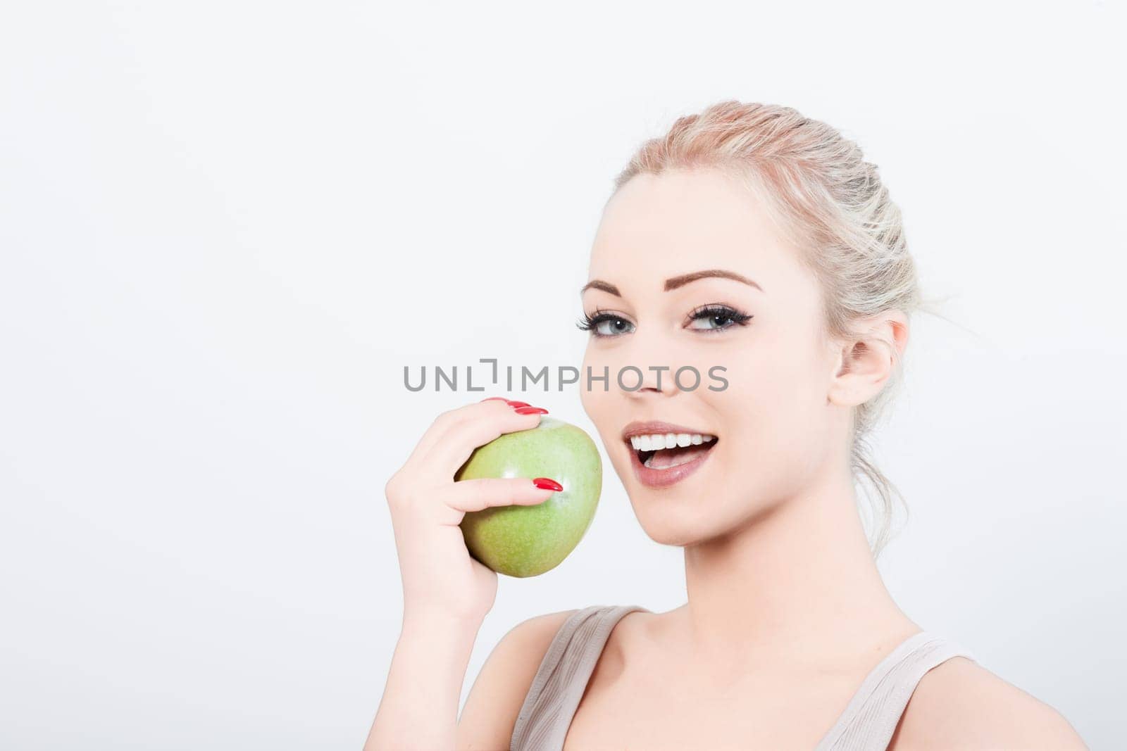 Cheerful woman with blonde hair tied back bites a fresh green apple against a white background, showcasing a healthy lifestyle with a bright smile