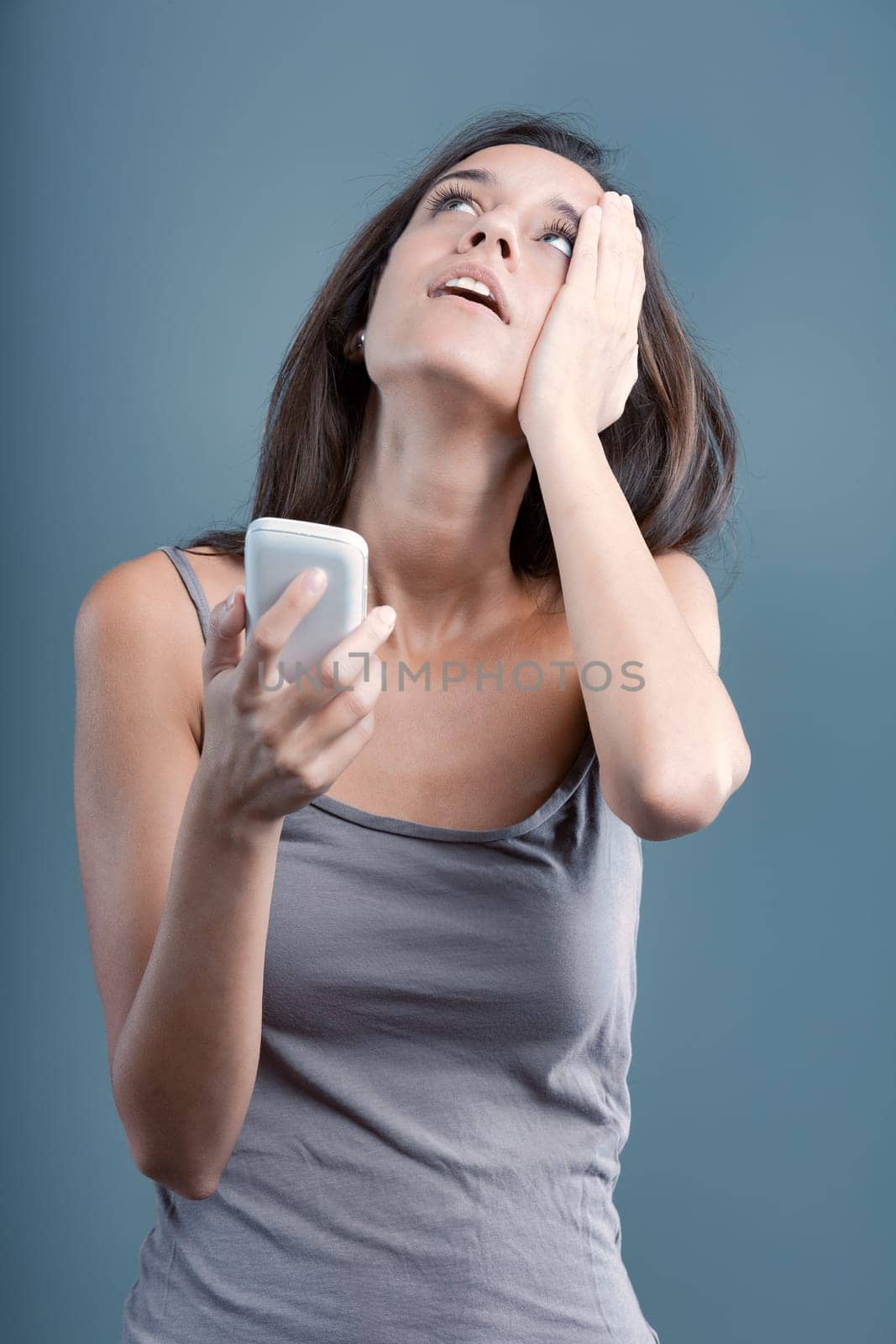 Stressed woman holding phone indoors against blue and gray background, looking up with frustration and concern, facing communication issues