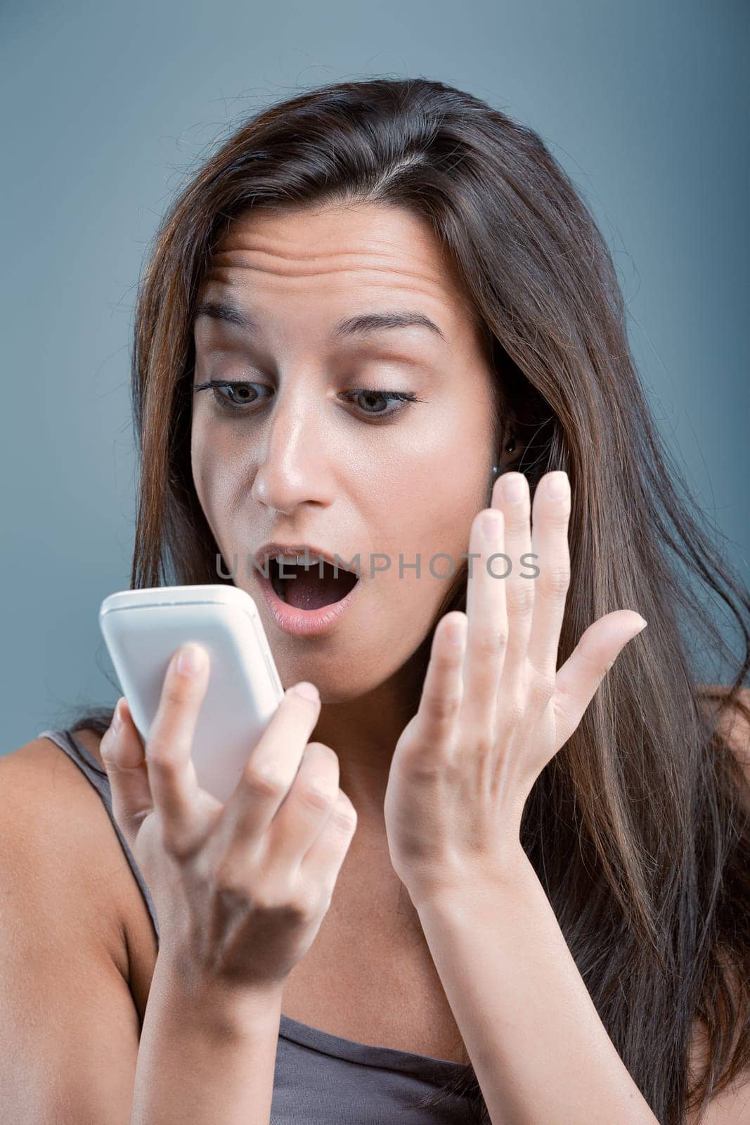 Surprised woman holding smartphone, reacting to shocking content, against blue background. Expression conveys disbelief and wonder. Browsing internet, amazed by message