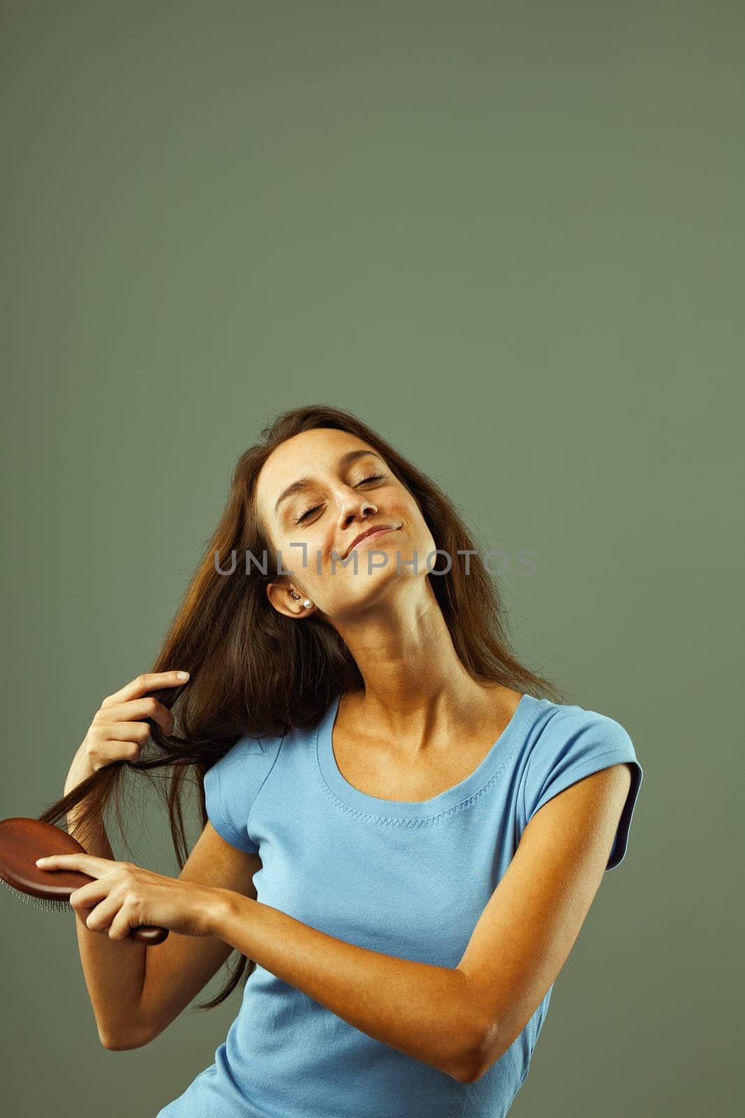Young woman with long, dark hair happily brushes her hair with a wooden hairbrush, eyes closed, smiling serenely, showcasing her healthy hair care routine against a green background