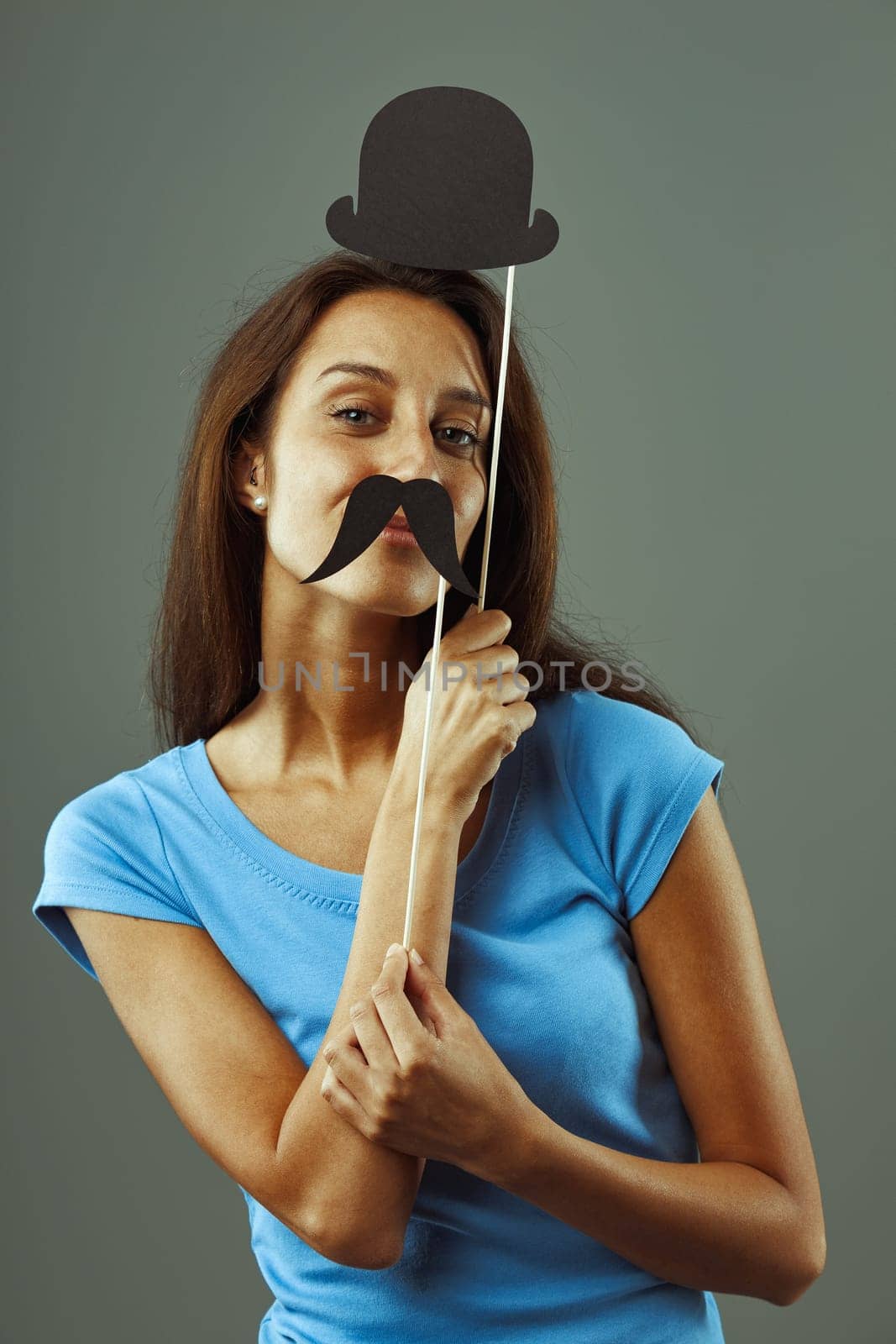 Young woman poses playfully with a fake mustache and bowler hat, radiating joy and happiness against a grey background