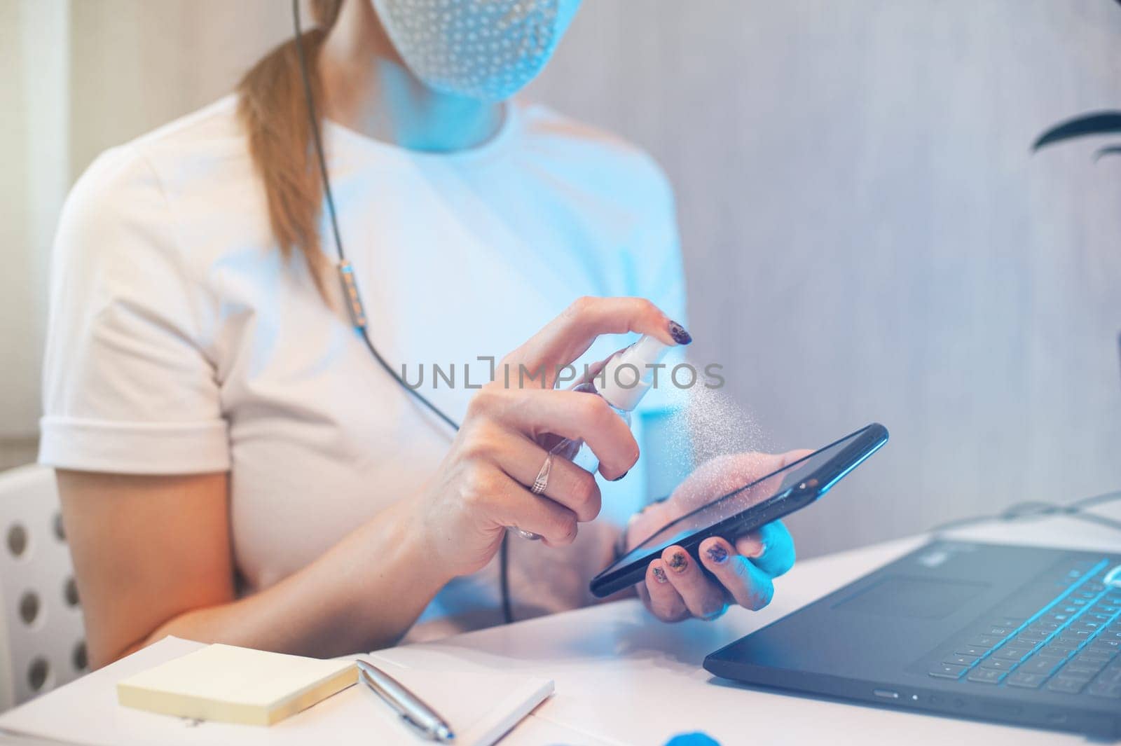 Woman disinfects the surface of the phone by sanitizer spray on the working place. Coronavirus concept. Woman in quarantine for coronavirus covid-19 working from home.