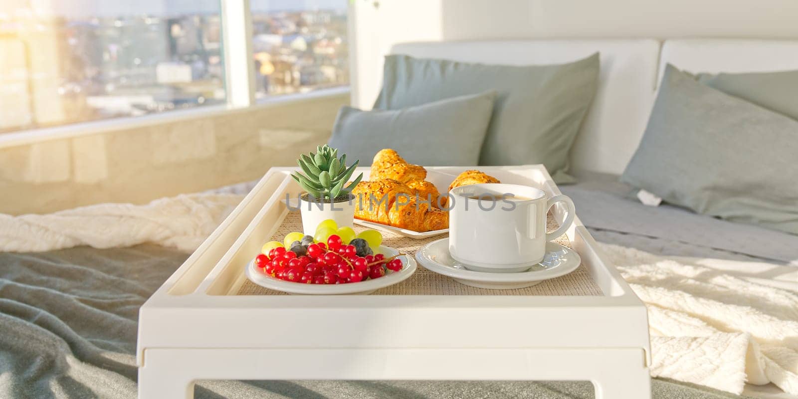 White Tray with tasty breakfast on bed in light bedroom with big windows in skyscraper. white table with tasty breakfast in bed at bed room in skyscraper