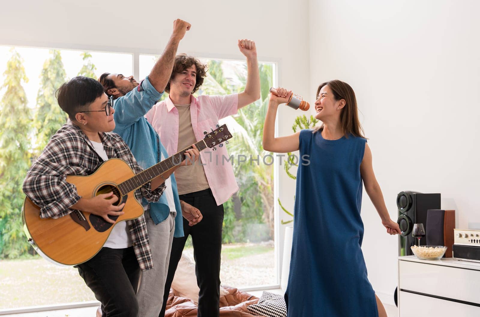 Group of friends having fun at home. One playing guitar, while others singing and cheering with raised hands. Multicultural people lively and joyful party together on weekend.