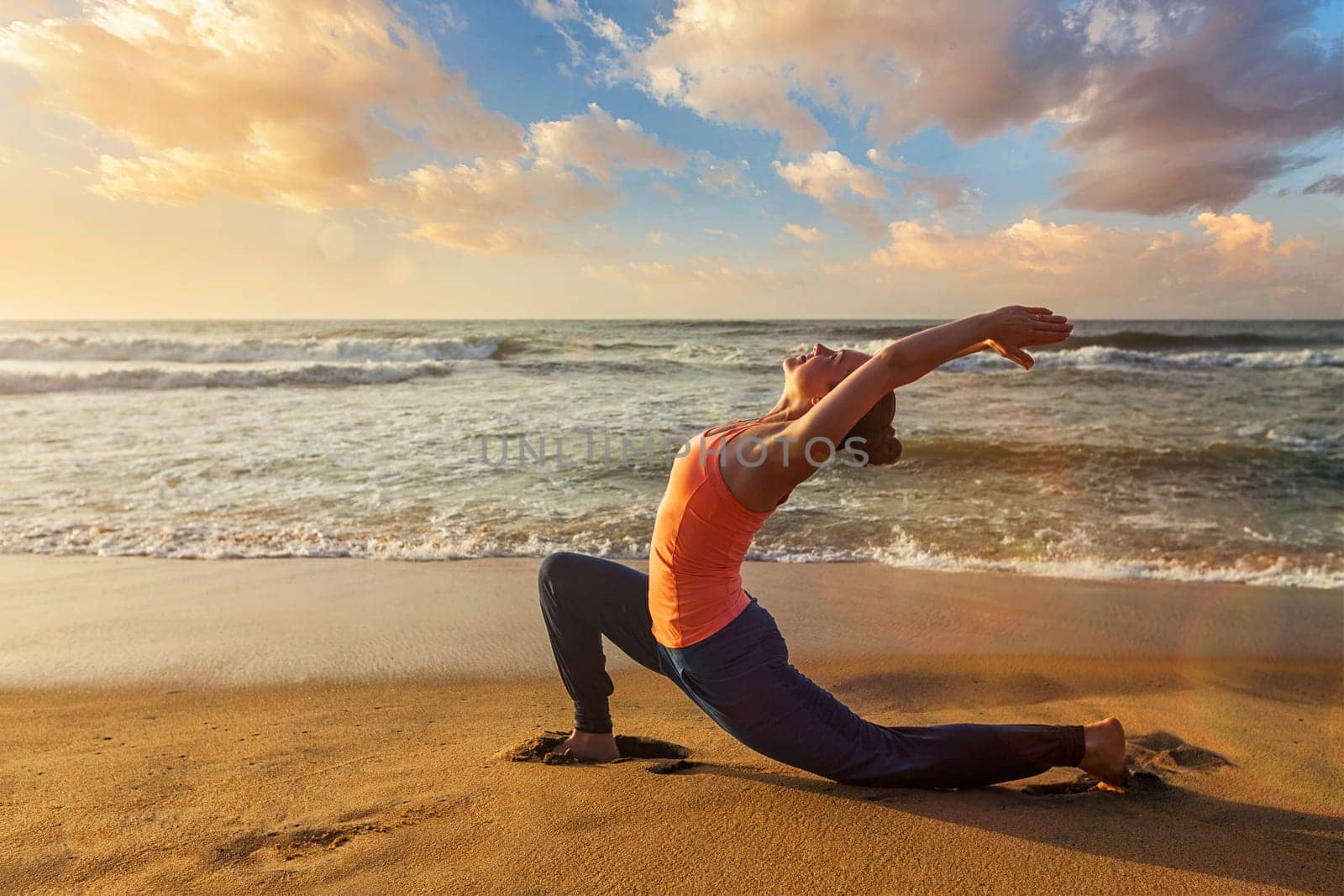 Yoga outdoors - sporty fit woman practices yoga Anjaneyasana - low crescent lunge pose outdoors at beach on sunset