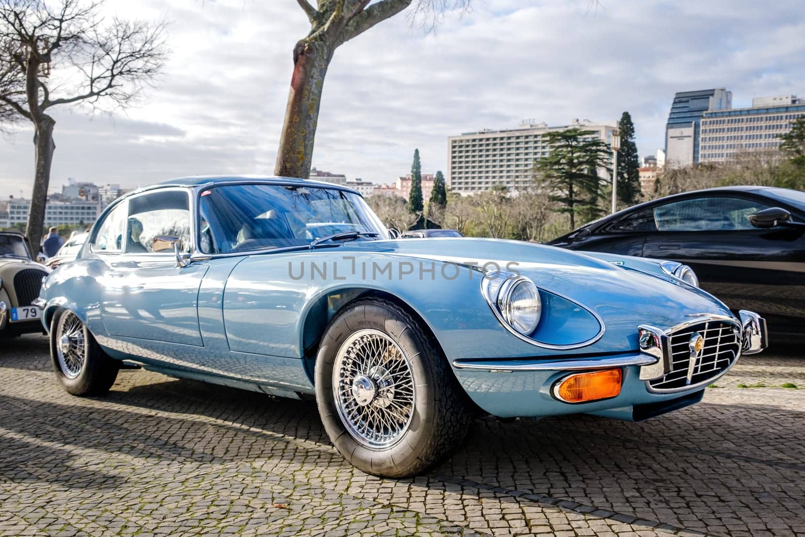 Lisbon, Portugal - Jan 20, 2024: A classic light-blue E-Type Jaguar Series 3 coupe car with chrome detailing parked on a cobblestone path near green park.