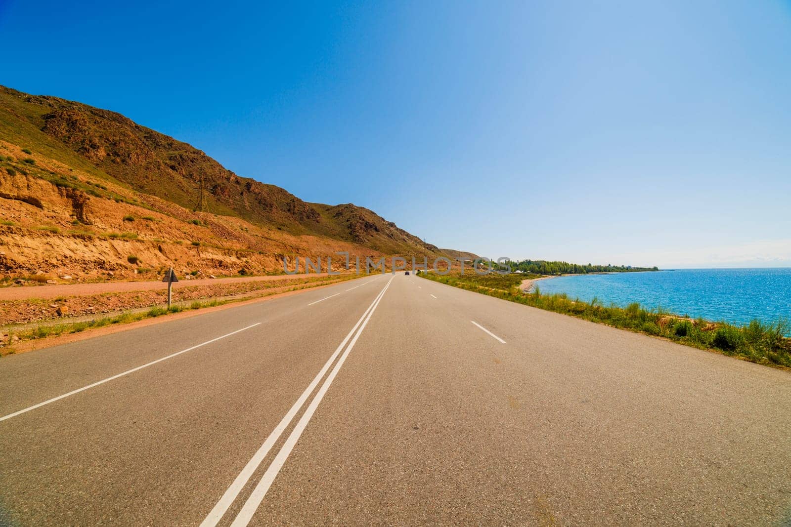 asphalt interstate road between yellow sedimentary rock mountain and large lake at sunny summer day, wide angle view