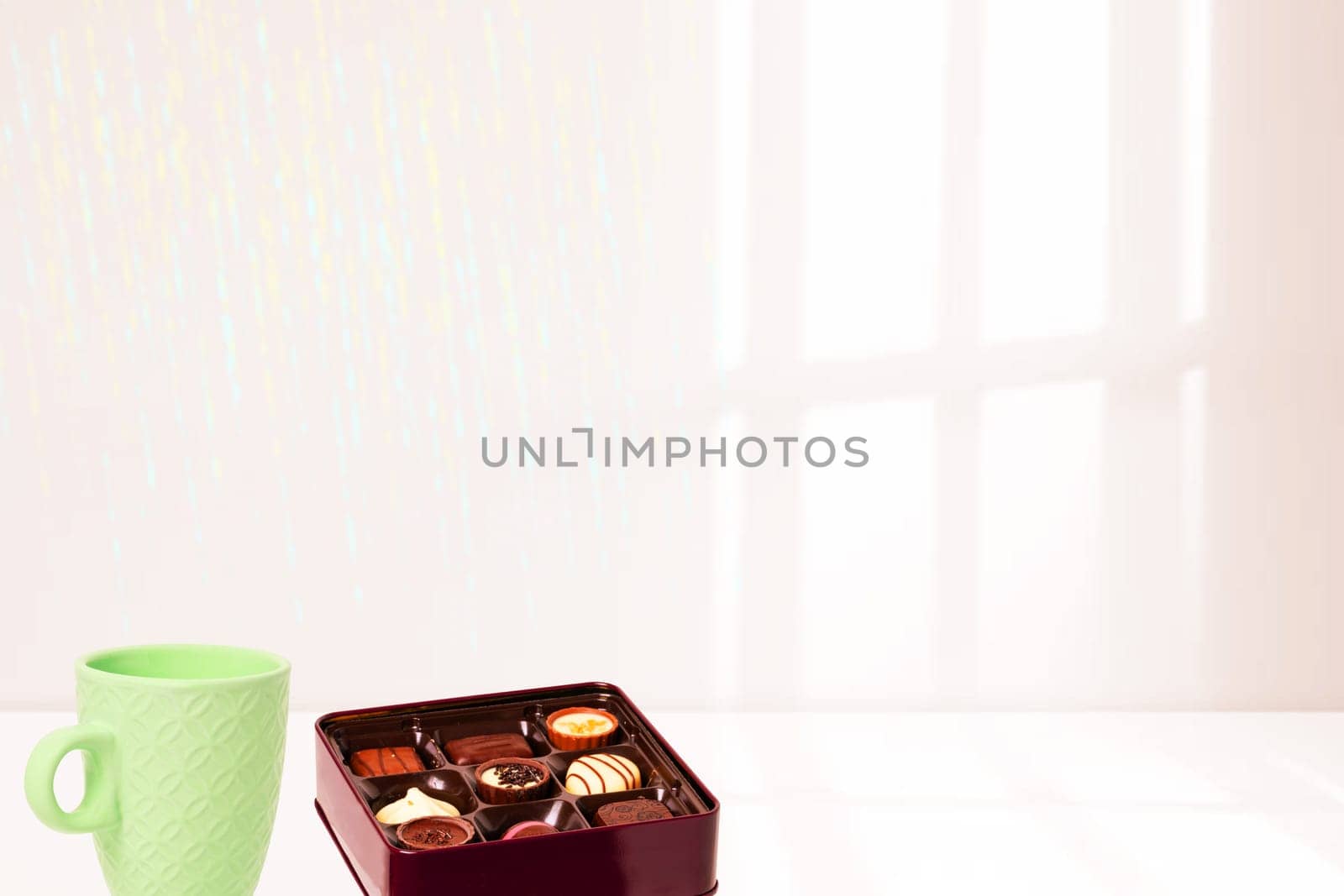 A box of fine chocolate pralines and a cup of coffee on a table against blurred curtain window background. Template for your product display montage. Space for design.