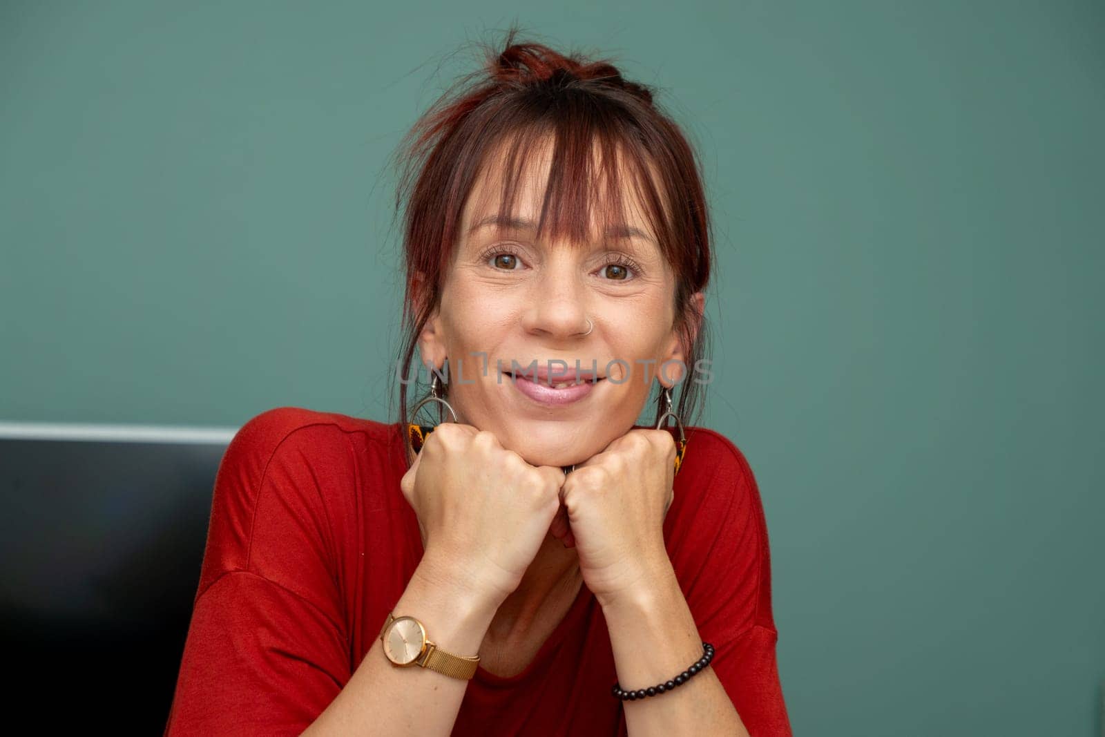 A woman with red hair and a red shirt is smiling and holding her hands together. She is wearing a watch and a bracelet