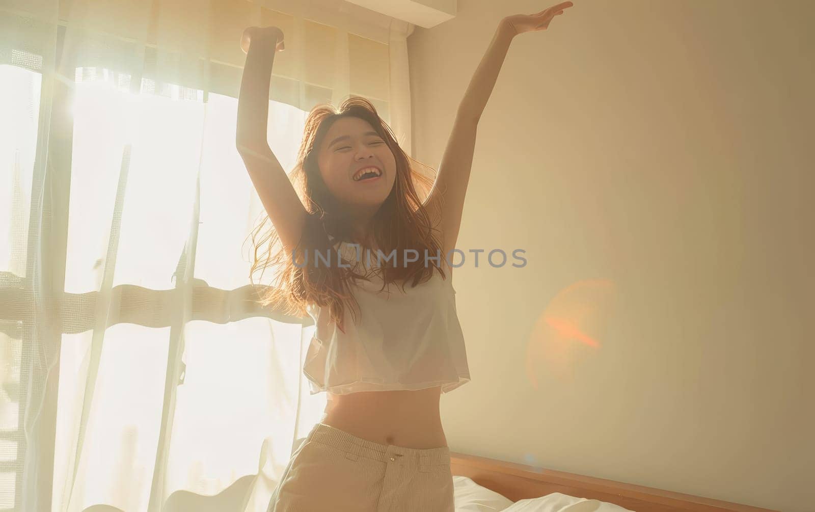 A young woman in a sunlit room stretches her arms in exultation, her silhouette glowing against the bright morning light.
