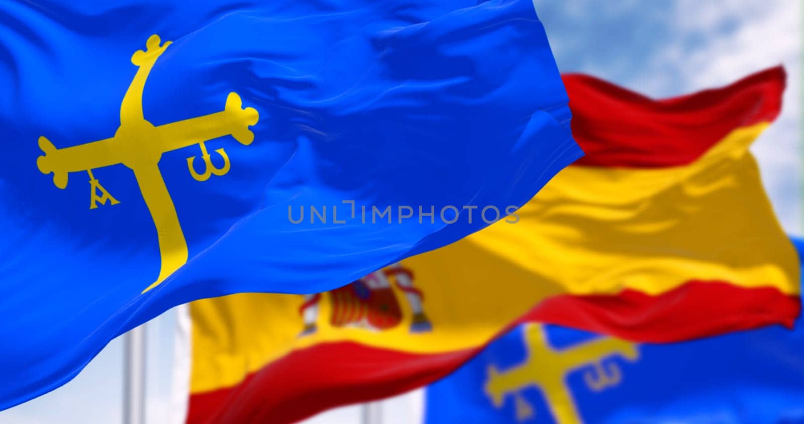 The Asturias flags waving with Spanish flag on a clear day. Autonomous community in northern Spain. Blue with golden victory cross. 3d illustration render. Rippling fabric. Selective focus