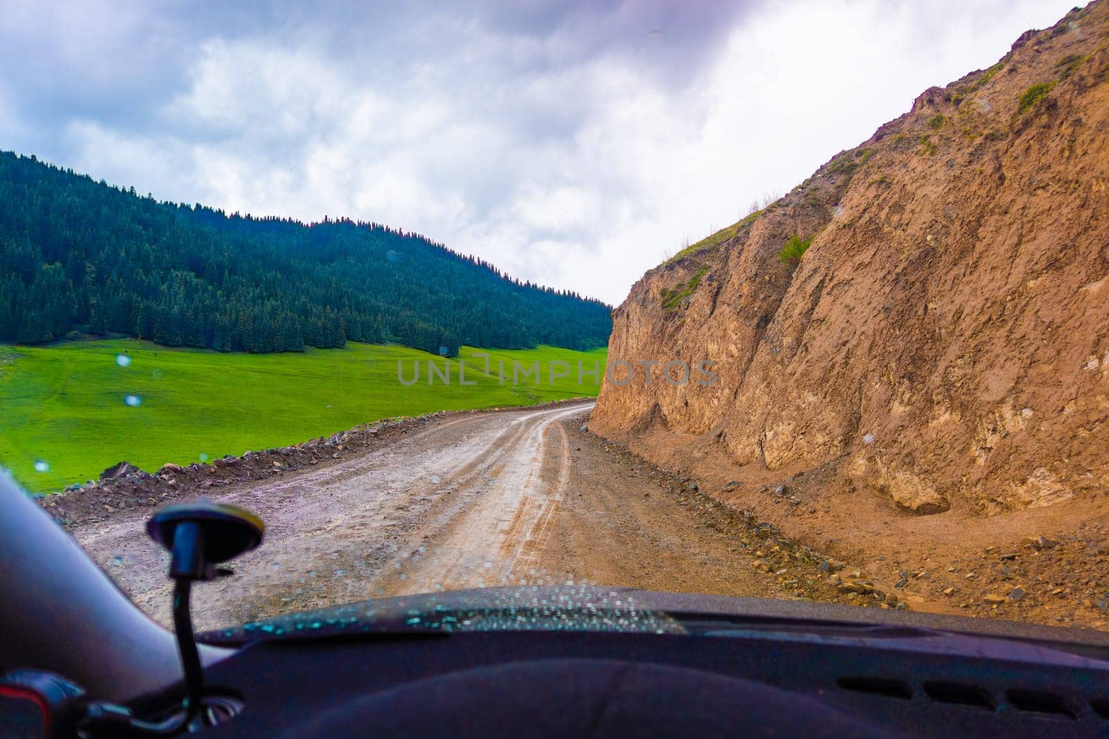 driving along the dirt road at rainy summer day in mountains with spruce forest on mountain slope, view from driver perspective