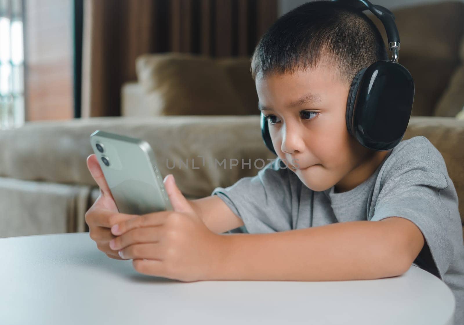 Cute elementary school boy wearing headphones holding a smartphone. Boy studying online on smartphone or homeschooling, listening to music or playing games, Concept of online learning via smartphone.