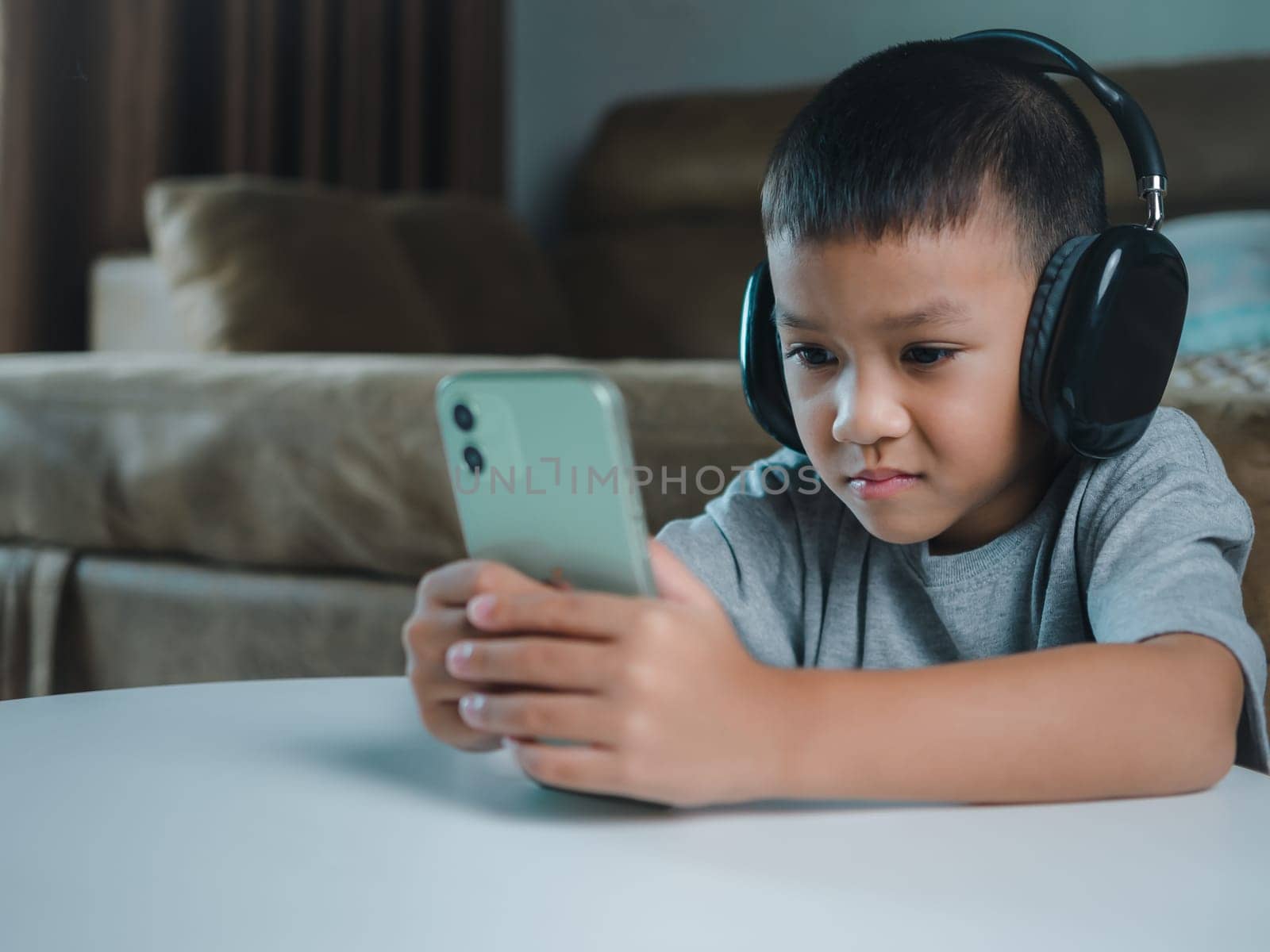 Cute elementary school boy wearing headphones holding a smartphone. Boy studying online on smartphone or homeschooling, listening to music or playing games, Concept of online learning via smartphone.