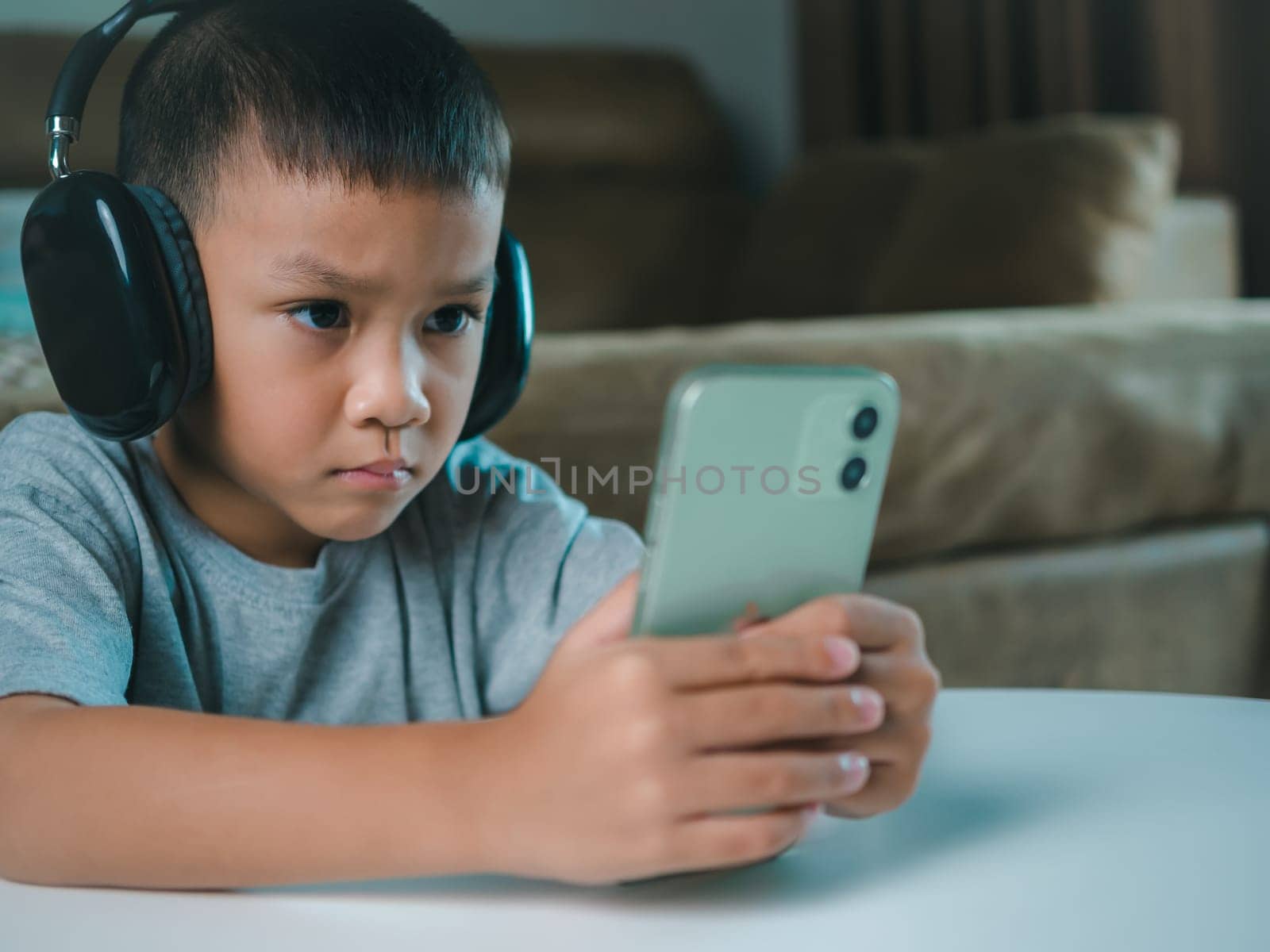 Cute elementary school boy wearing headphones holding a smartphone. Boy studying online on smartphone or homeschooling, listening to music or playing games, Concept of online learning via smartphone.