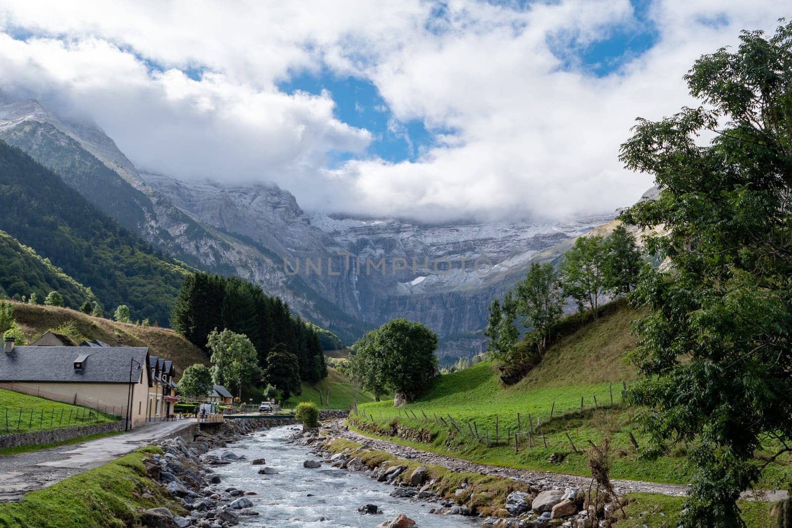 The Cirque of Gavarnie, french Pyrenes mountains. The mountains are covered in snow and the sky is cloudy. The river is surrounded by a lush green valley