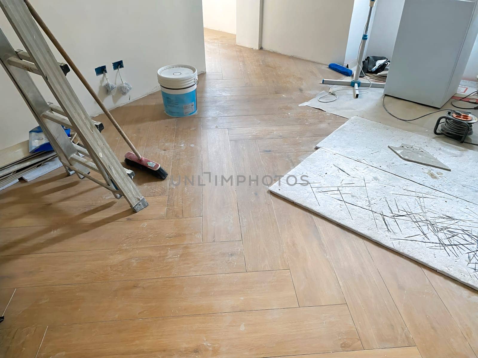 Indoor view of a room under renovation, featuring newly installed wooden flooring, a ladder, a broom, and various construction tools