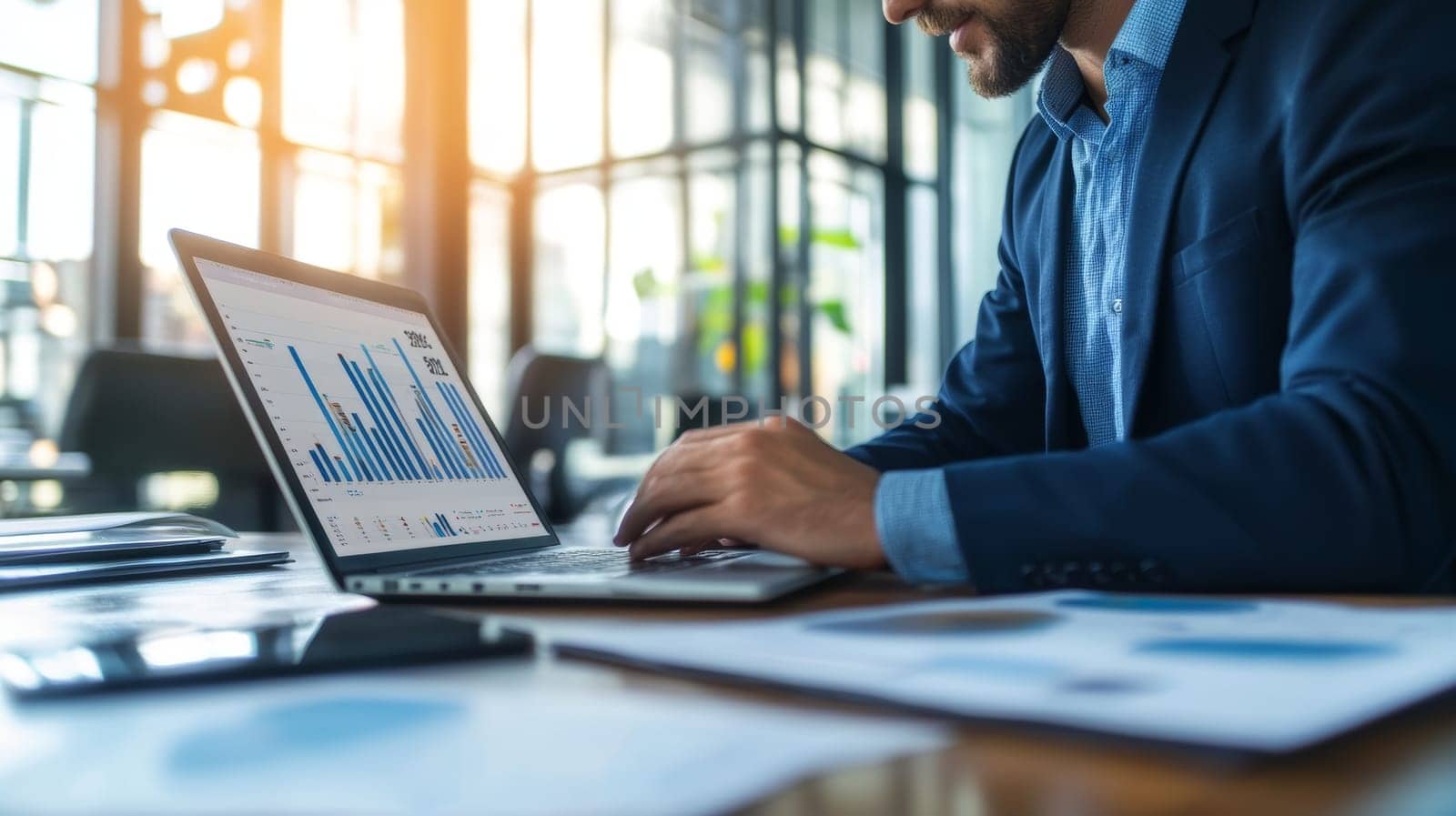 Bearded businessman in blue shirt examining financial charts and graphs on laptop screen in modern office with sunlit windows.