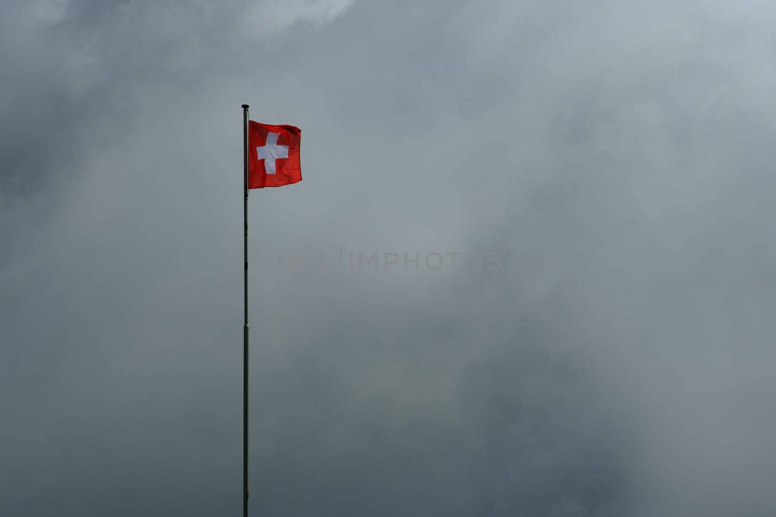 Swiss flag on flagpole waving in the wind against a cloudy background