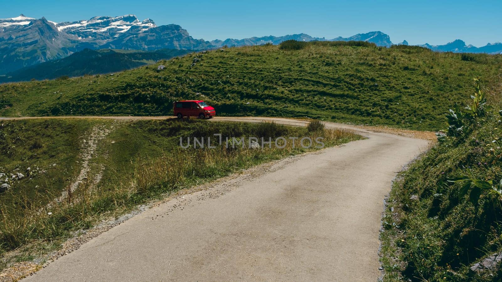 Red van driving uphill on a narrow alpine mountain road high up in the Vaud Alps on a bright day in summer, near Leysin with the Les Diablerets mountain range in the background. Switzerland