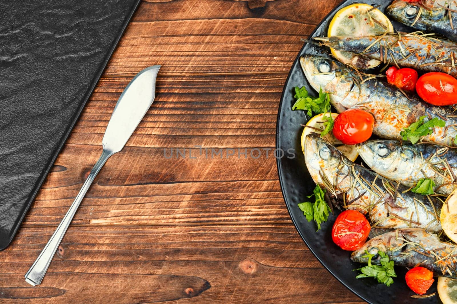 Baked sardine fish with lemon, cherry tomatoes and rosemary on ceramic plate on rustic wooden table. Healthy and tasty dinner. Copy space.