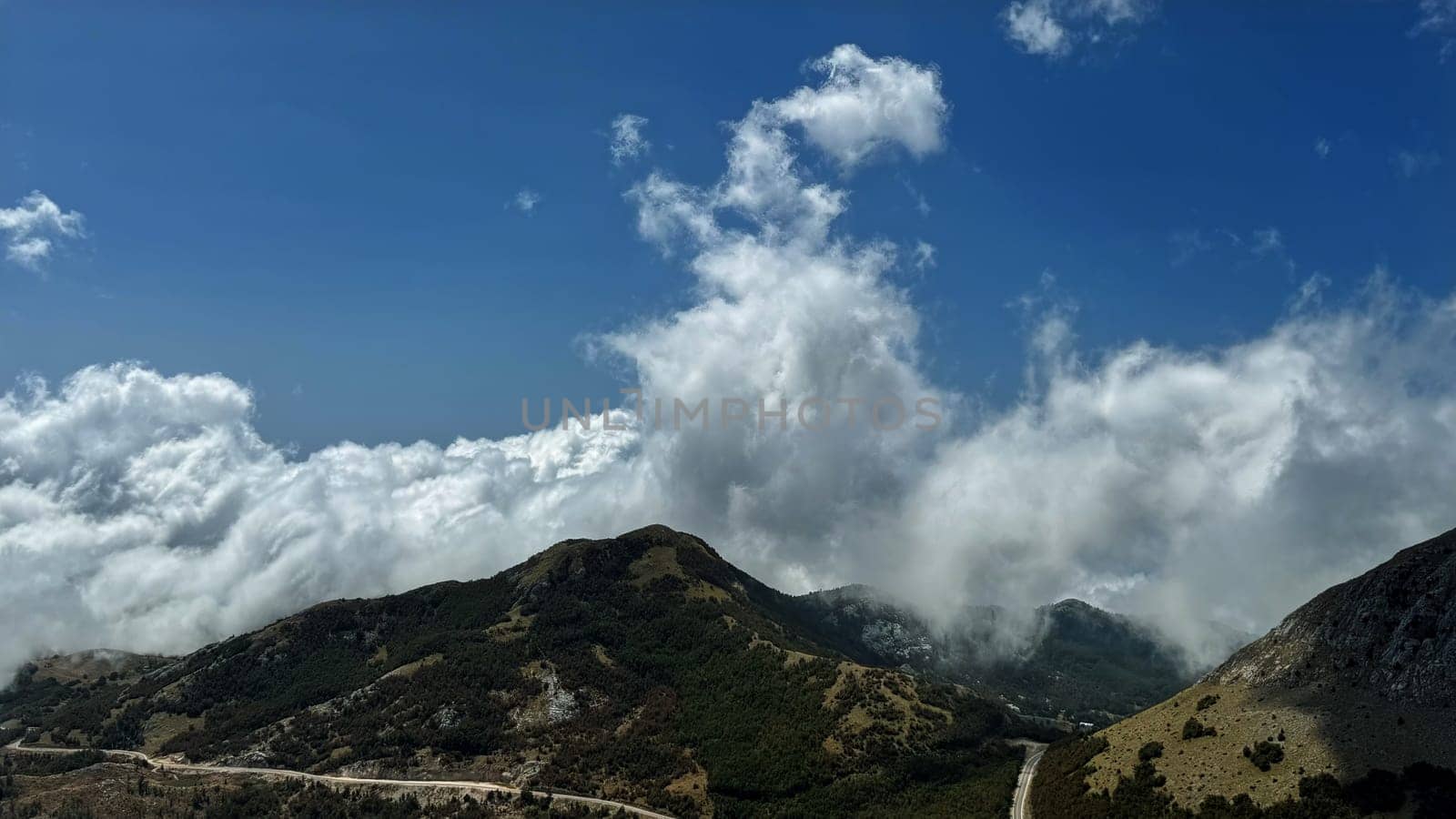 A captivating view of clouds rolling over a lush mountain range, with a winding road cutting through the green hills, under a bright blue sky.