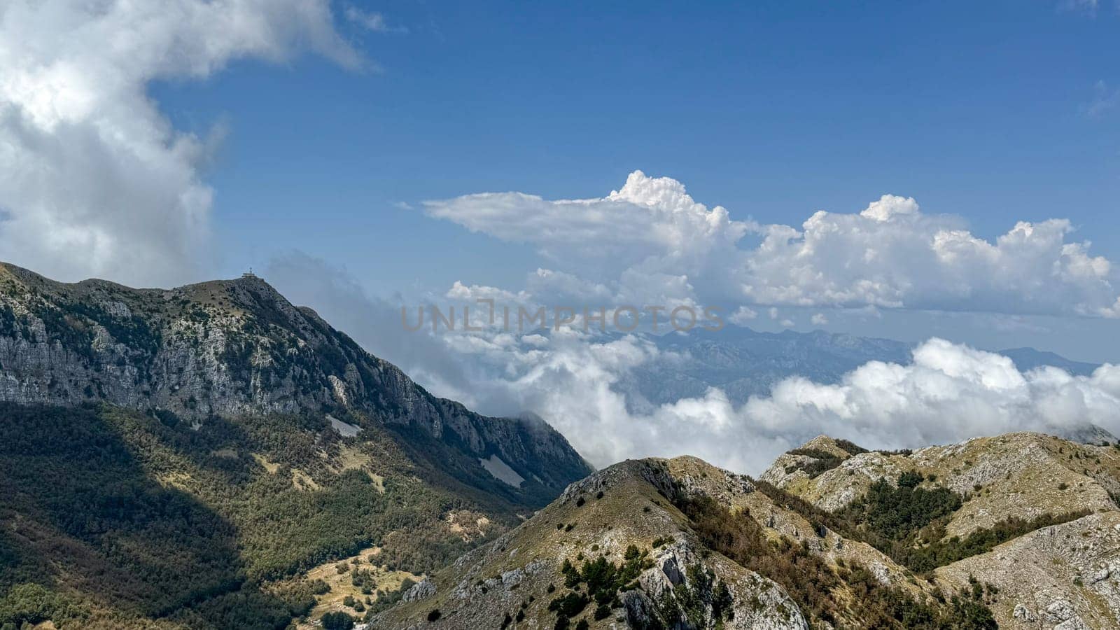A wide view of a majestic mountain range with rocky peaks and lush valleys, partially covered by clouds under a bright blue sky.