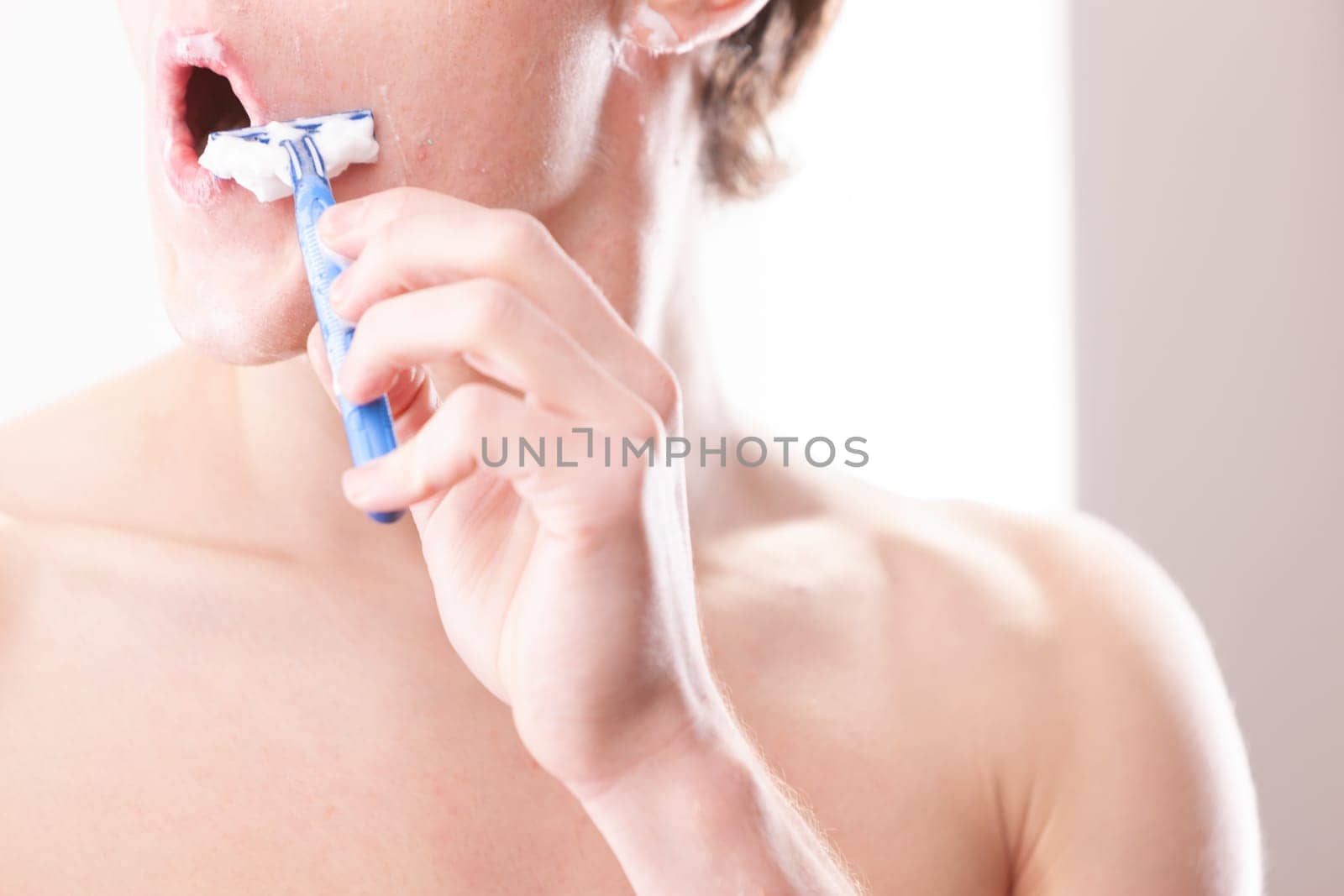 Young man is carefully shaving his face with a razor in the bathroom, applying shaving cream for a smooth and comfortable experience