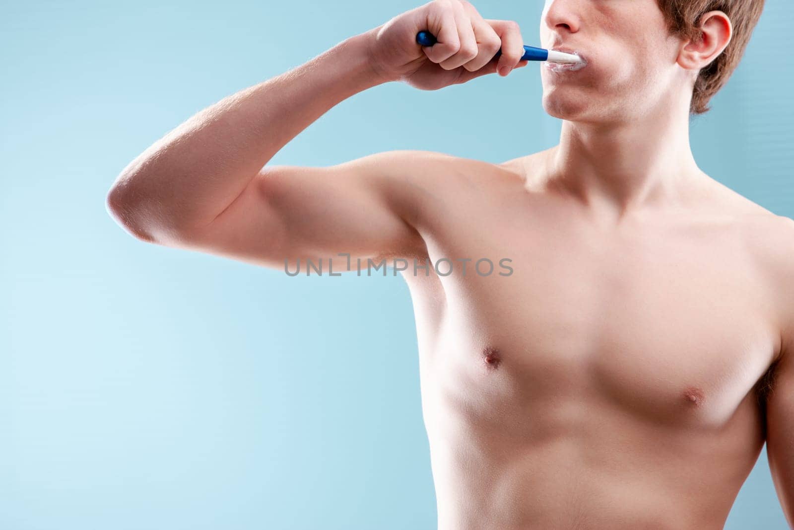 Young man brushing his teeth in the bathroom, focusing on oral hygiene and personal care with a fit body and a calming blue backdrop
