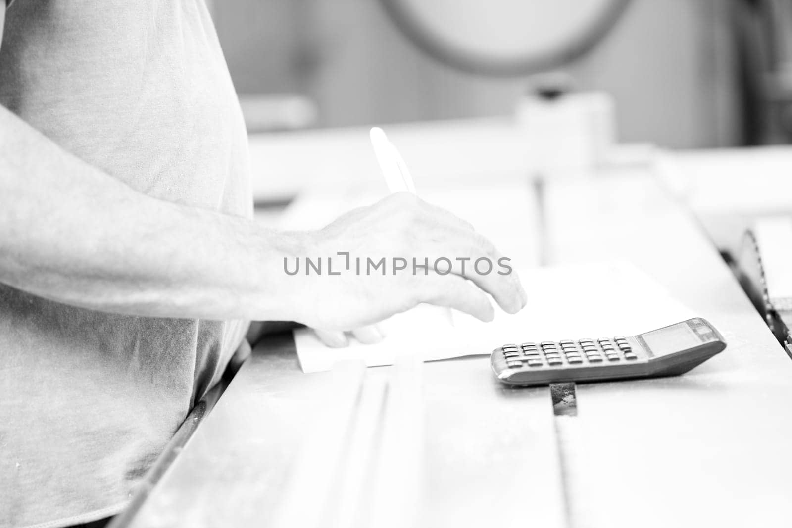 Carpenter is calculating costs in his workshop using a calculator and writing on a sheet of paper. - black n white -