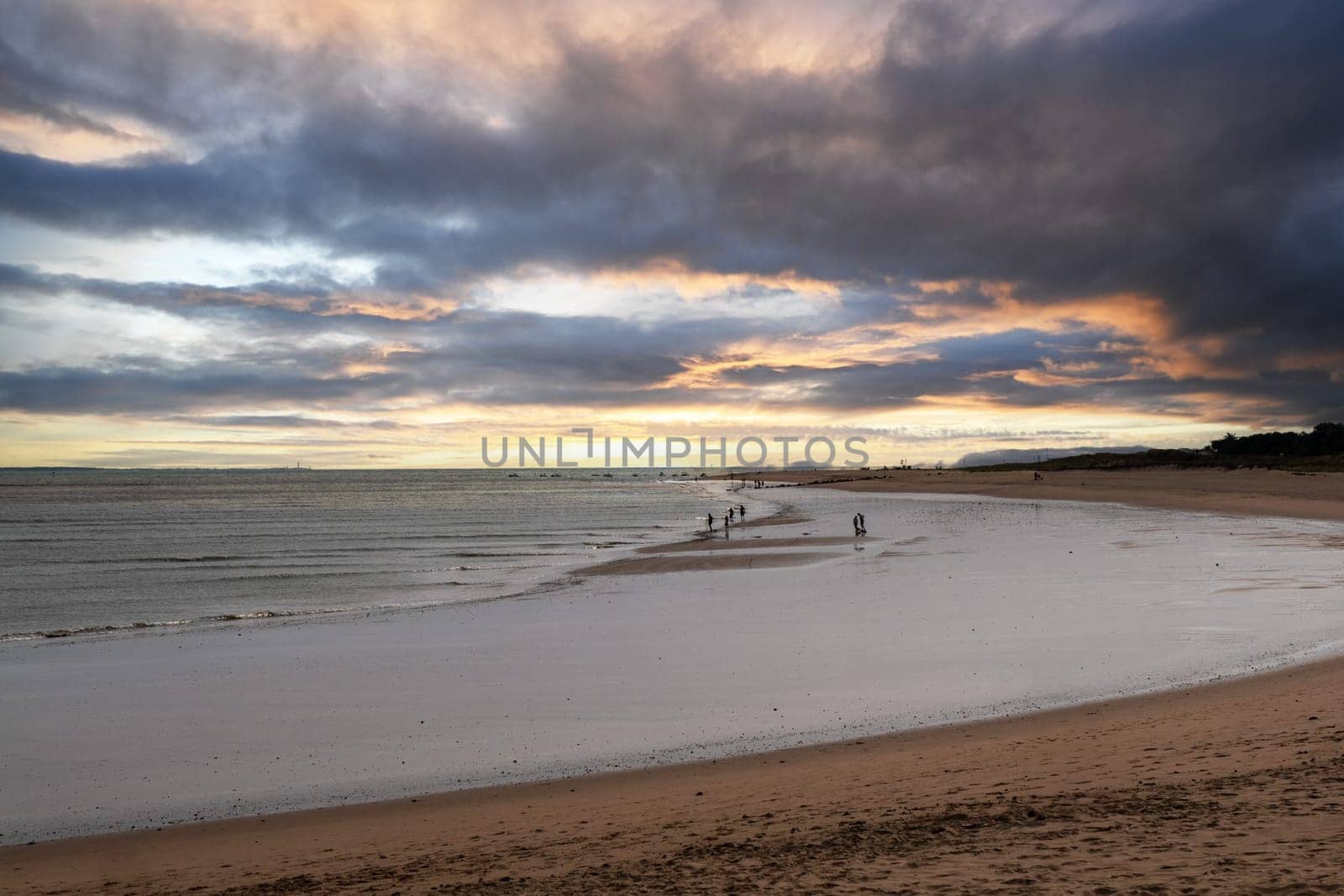 The beach is a serene and tranquil place, with a few people walking along the shoreline. The sky above is cloudy, creating a moody atmosphere. The water is calm, and the sand is soft and inviting